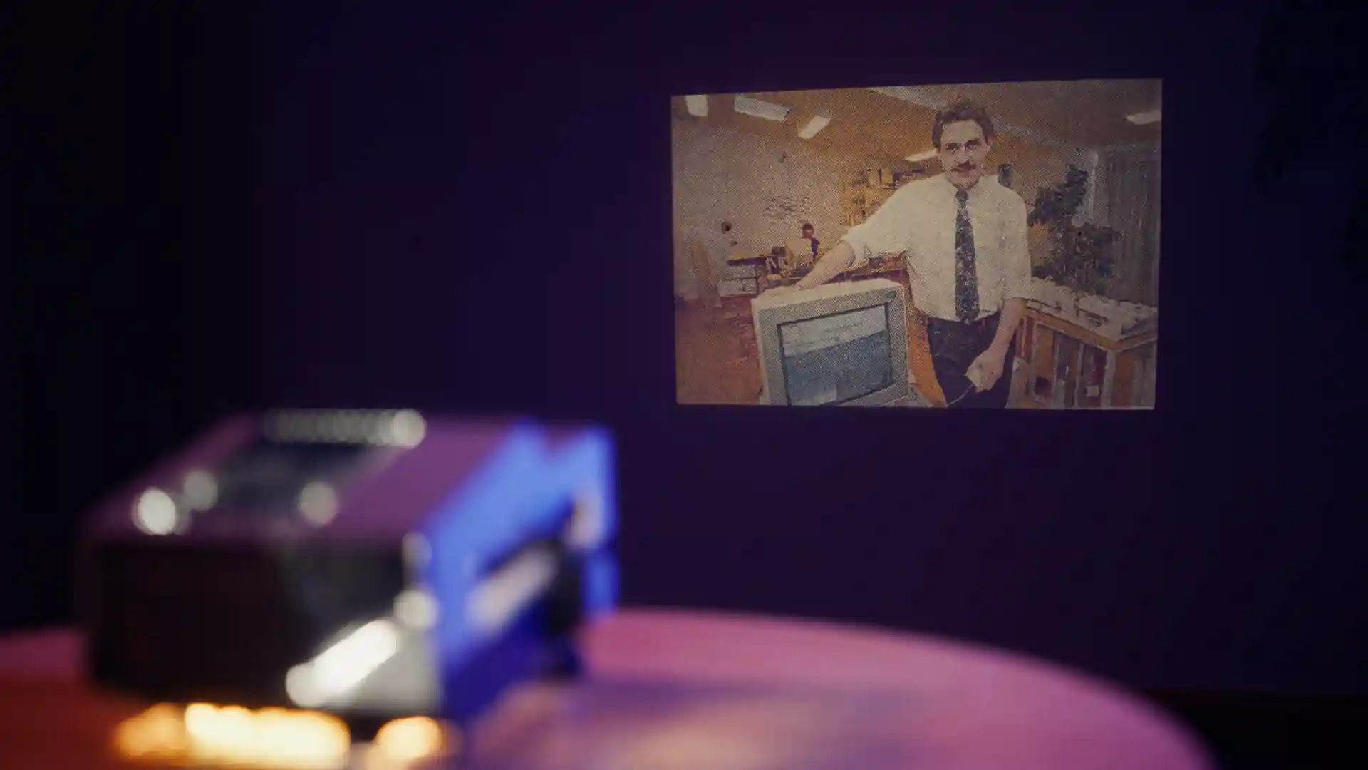 A slide projector displays a retro photo of a man in an office setting, pointing to an old computer monitor.