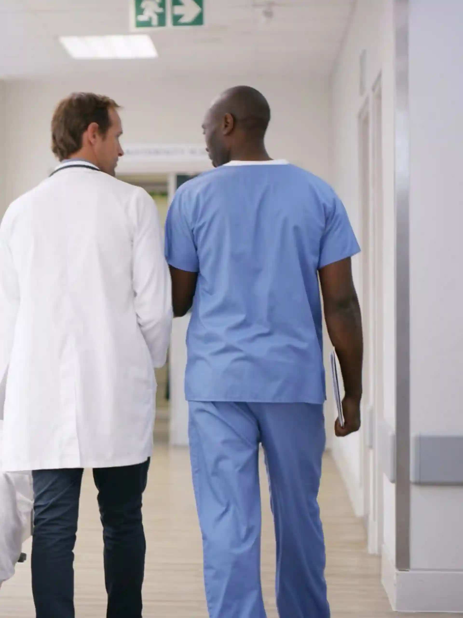 Healthcare team walking through a bright hospital corridor, including nurses in blue scrubs, a doctor in a white coat, and a nurse pushing a hospital bed.