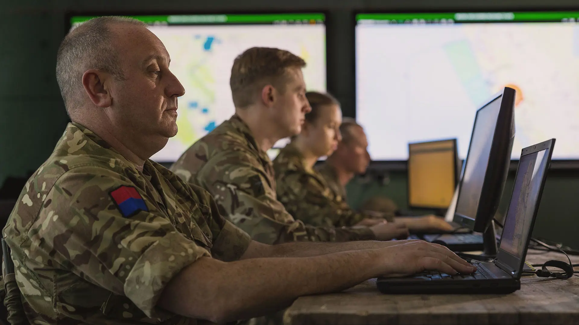 Military personnel in camouflage uniforms work on laptops inside a headquarters, monitoring digital maps and operational data displayed on large screens with SitaWare software.
