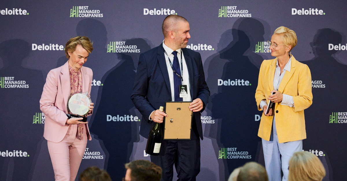 Tre personer står på scenen under Deloittes Best Managed Companies-prisutdeling og holder henholdsvis et trofé, et innrammet diplom og en flaske.
