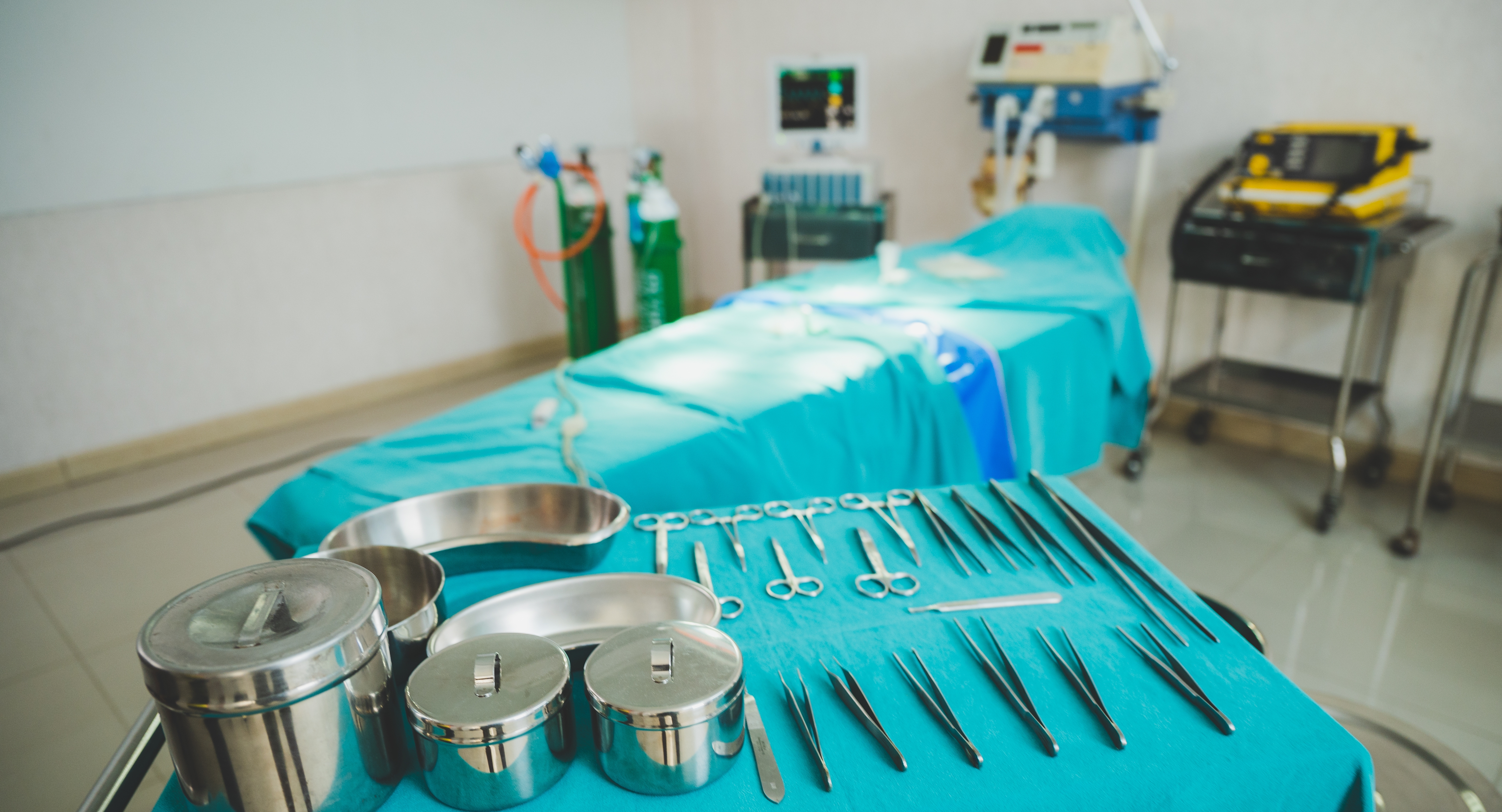 Surgical instruments laid out on a tray in an operating theatre, with an operating table and medical equipment in the background