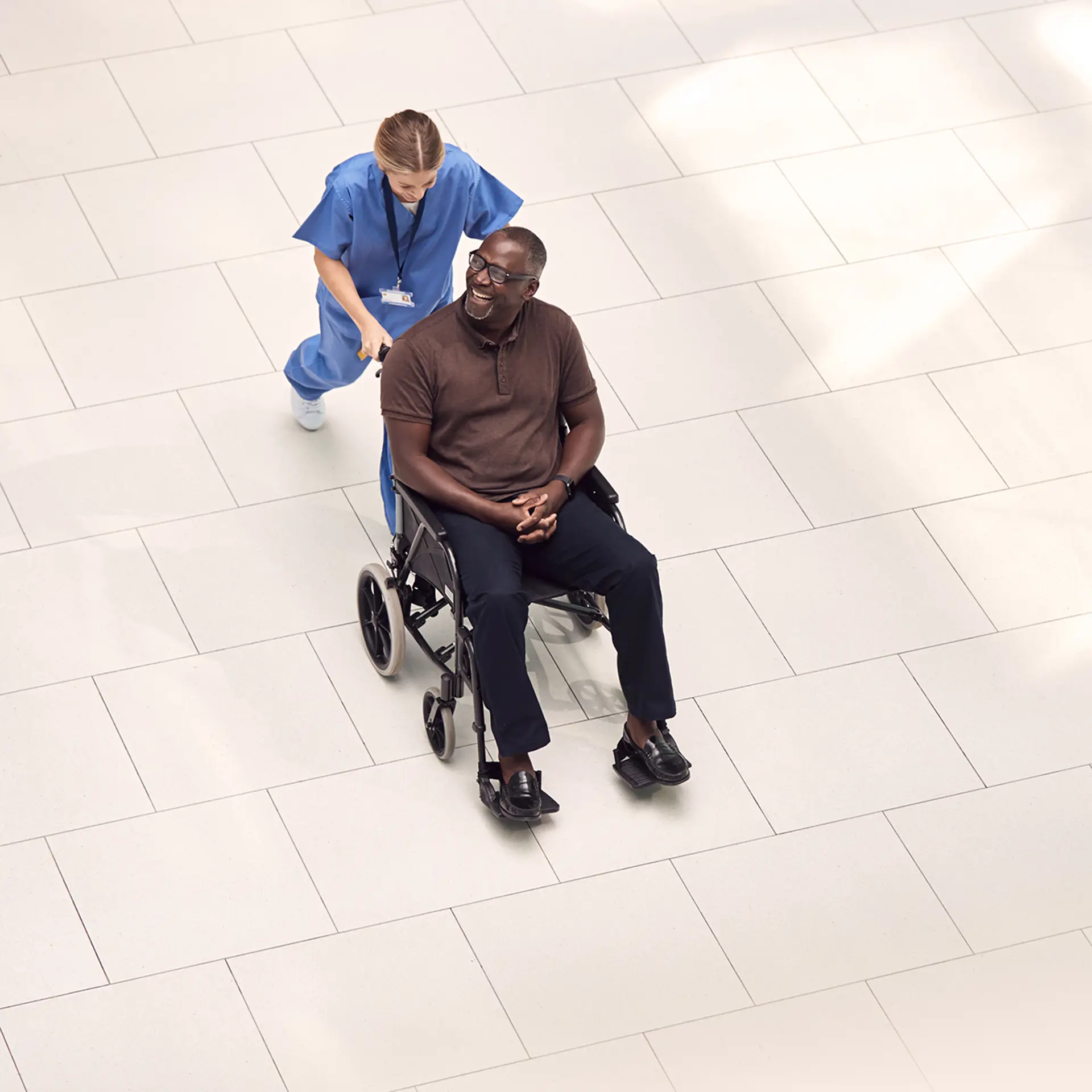 A healthcare worker in blue scrubs pushes an elderly man in a wheelchair down a bright, modern hallway.