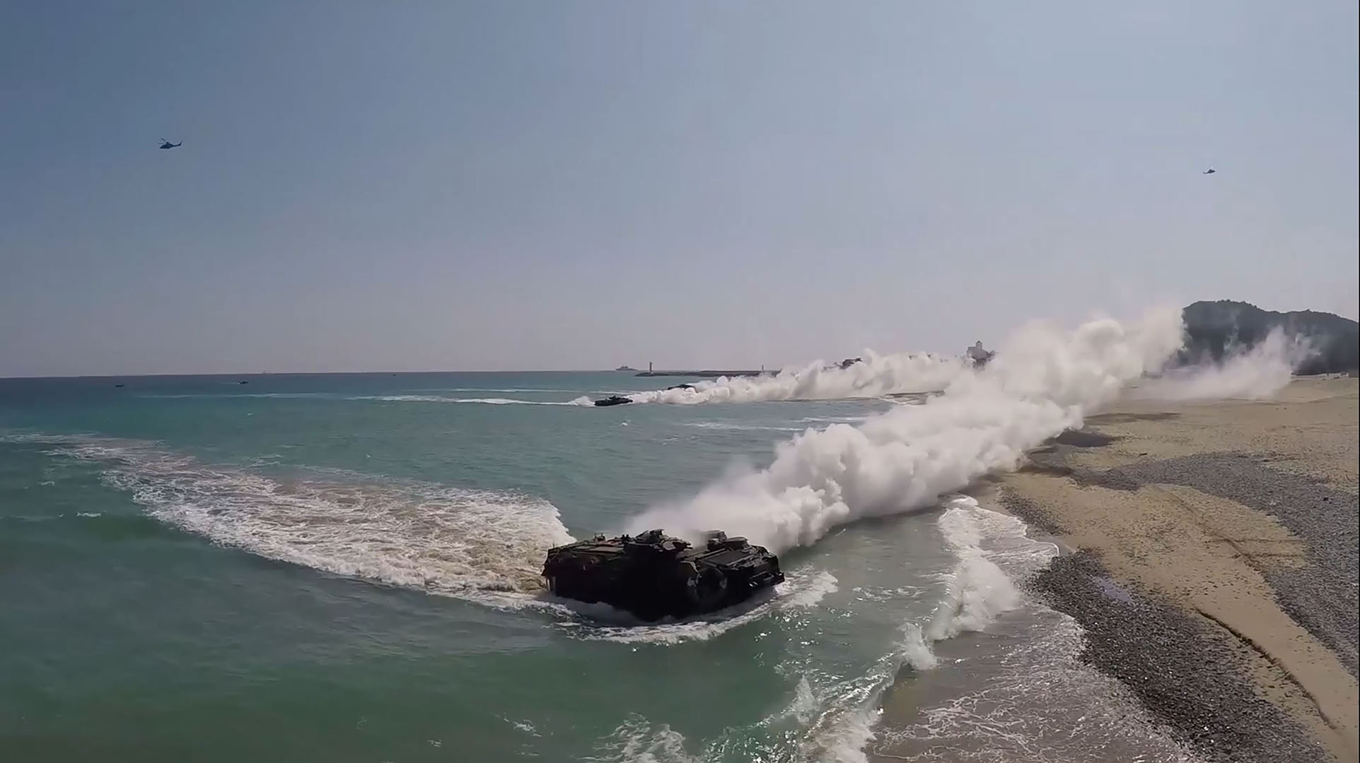 Amphibious armoured vehicles land on a beach, creating smoke trails, as helicopters fly overhead