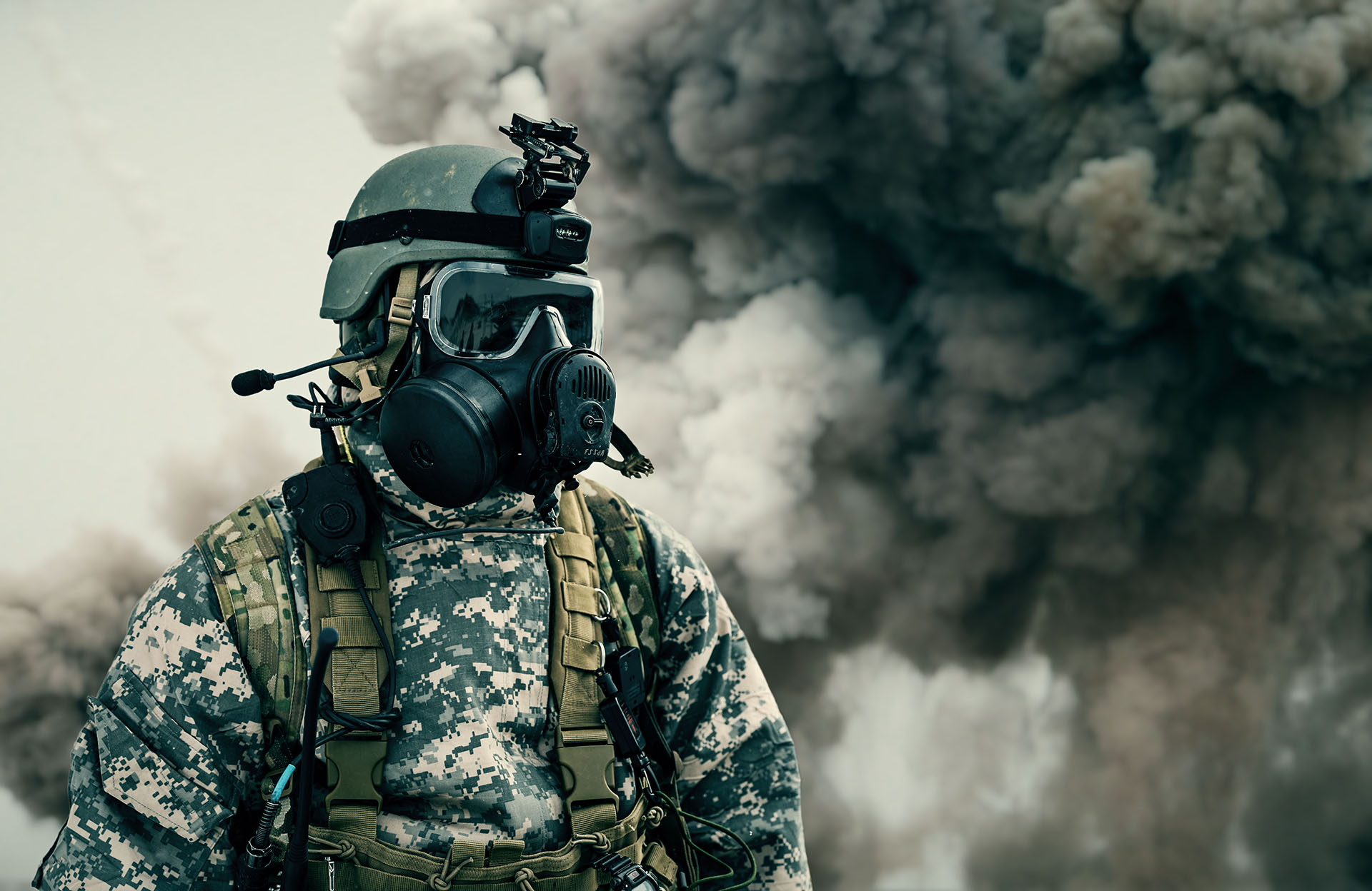 A soldier in CBRN protective gear with a gas mask, helmet and headset stands in front of thick, dark smoke.