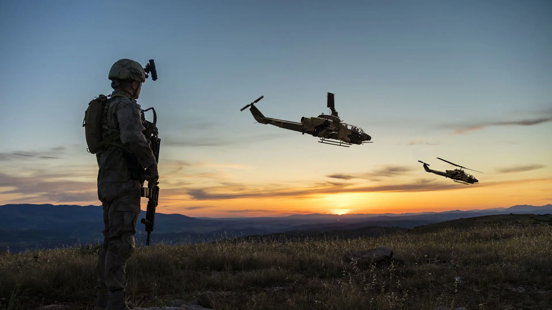 A soldier in tactical gear stands on a rocky hill at sunset, observing two military helicopters in flight