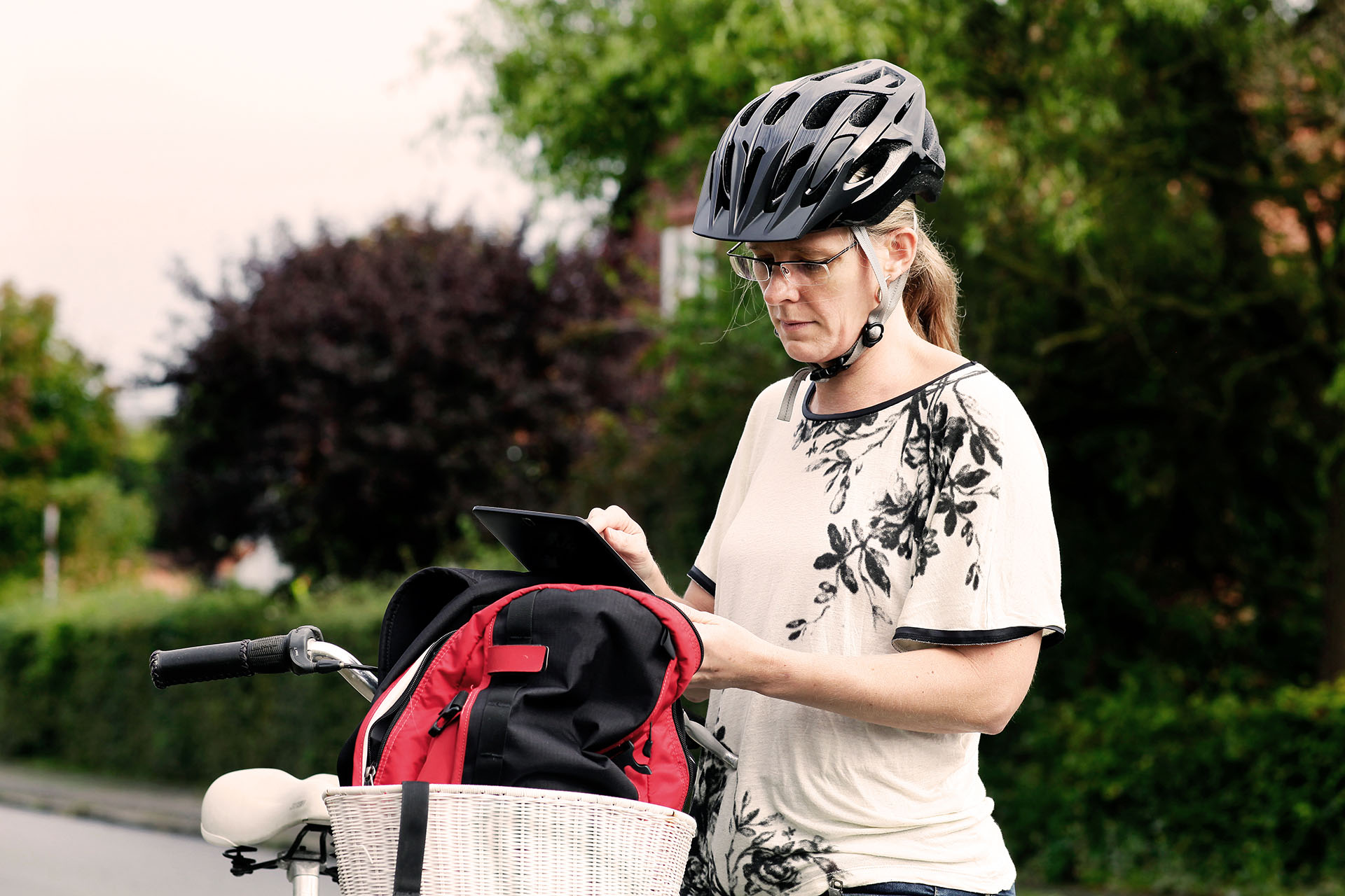 Sosialarbeider med sykkelhjelm bruker et nettbrett ved siden av sykkelen, med en rød og svart ryggsekk i sykkelkurven, i et boligområde.