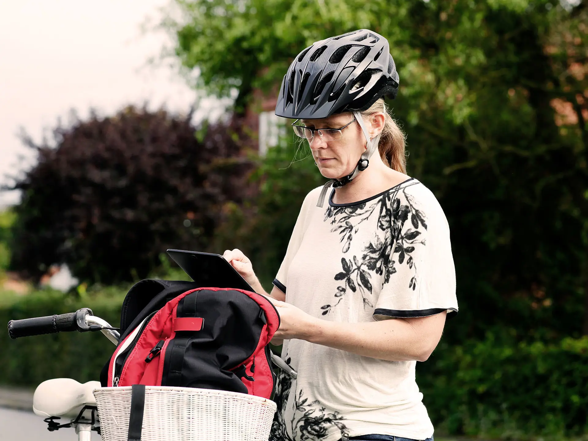 Sosialarbeider med sykkelhjelm bruker et nettbrett ved siden av sykkelen, med en rød og svart ryggsekk i sykkelkurven, i et boligområde.