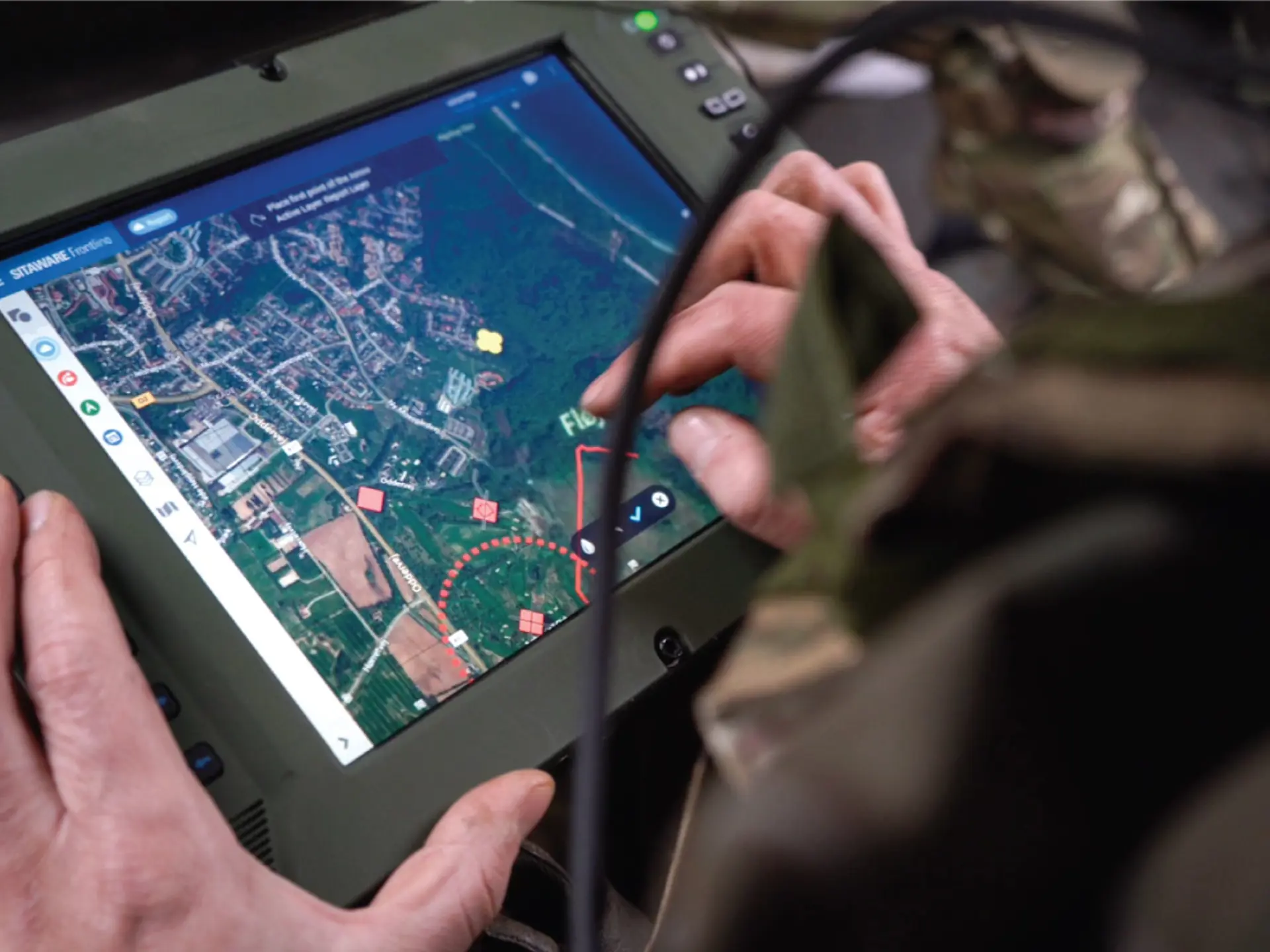 A soldier uses a rugged military tablet displaying a digital battlefield map with SitaWare software to coordinate frontline operations.
