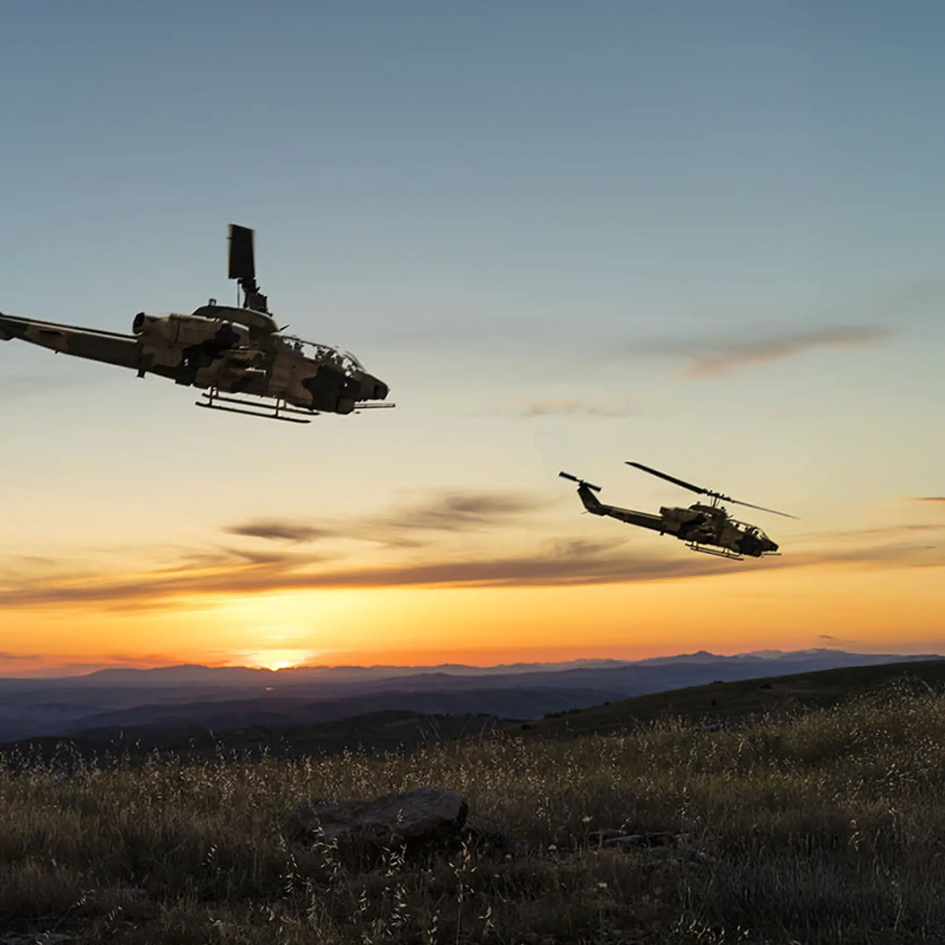 Soldier observing two military helicopters flying over a mountainous landscape at sunset, symbolizing real-time situational awareness enabled by SitaWare BattleCloud
