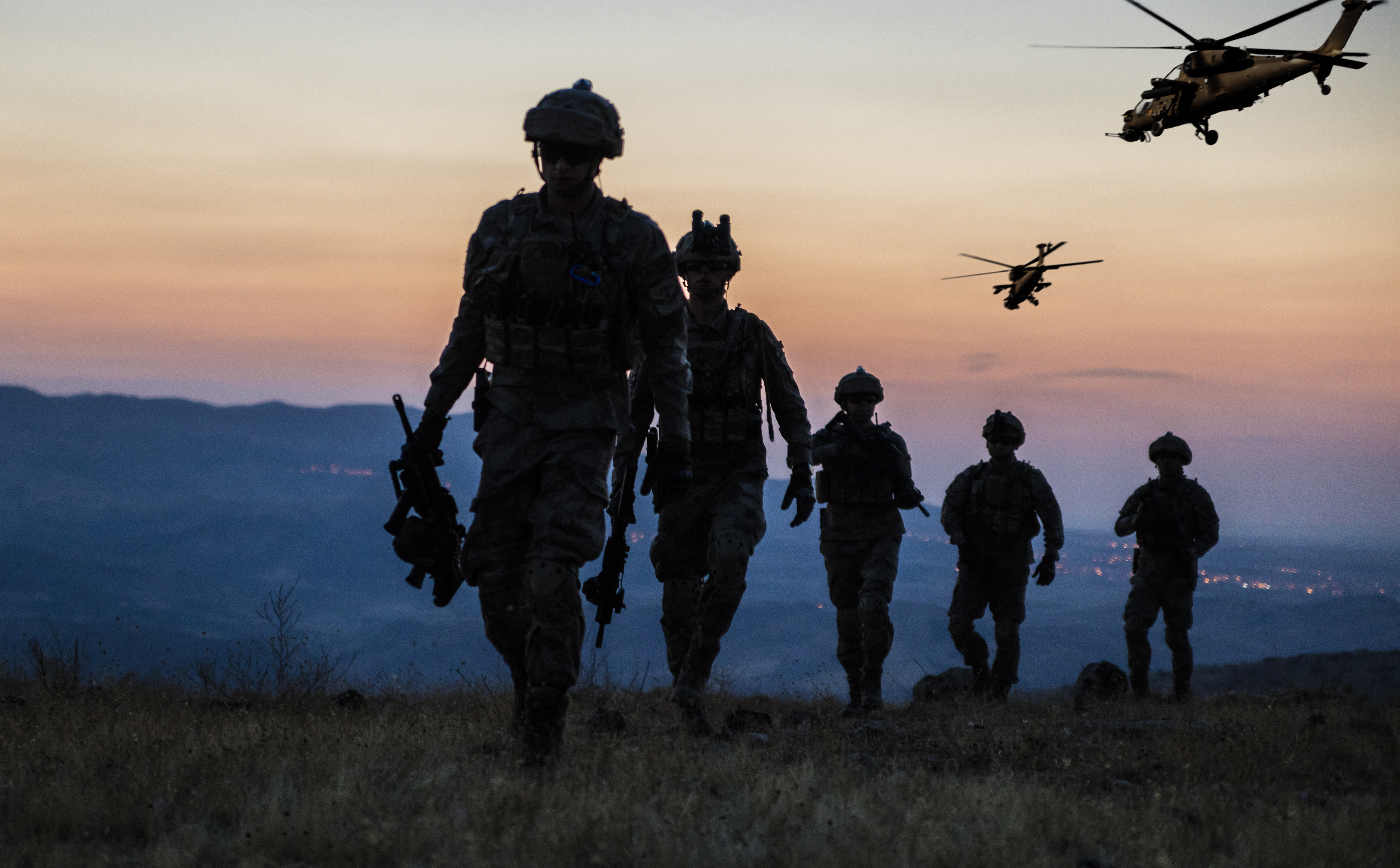Five soldiers with rifles walk across a grassy hilltop at dusk while two military helicopters fly over the valley behind them.