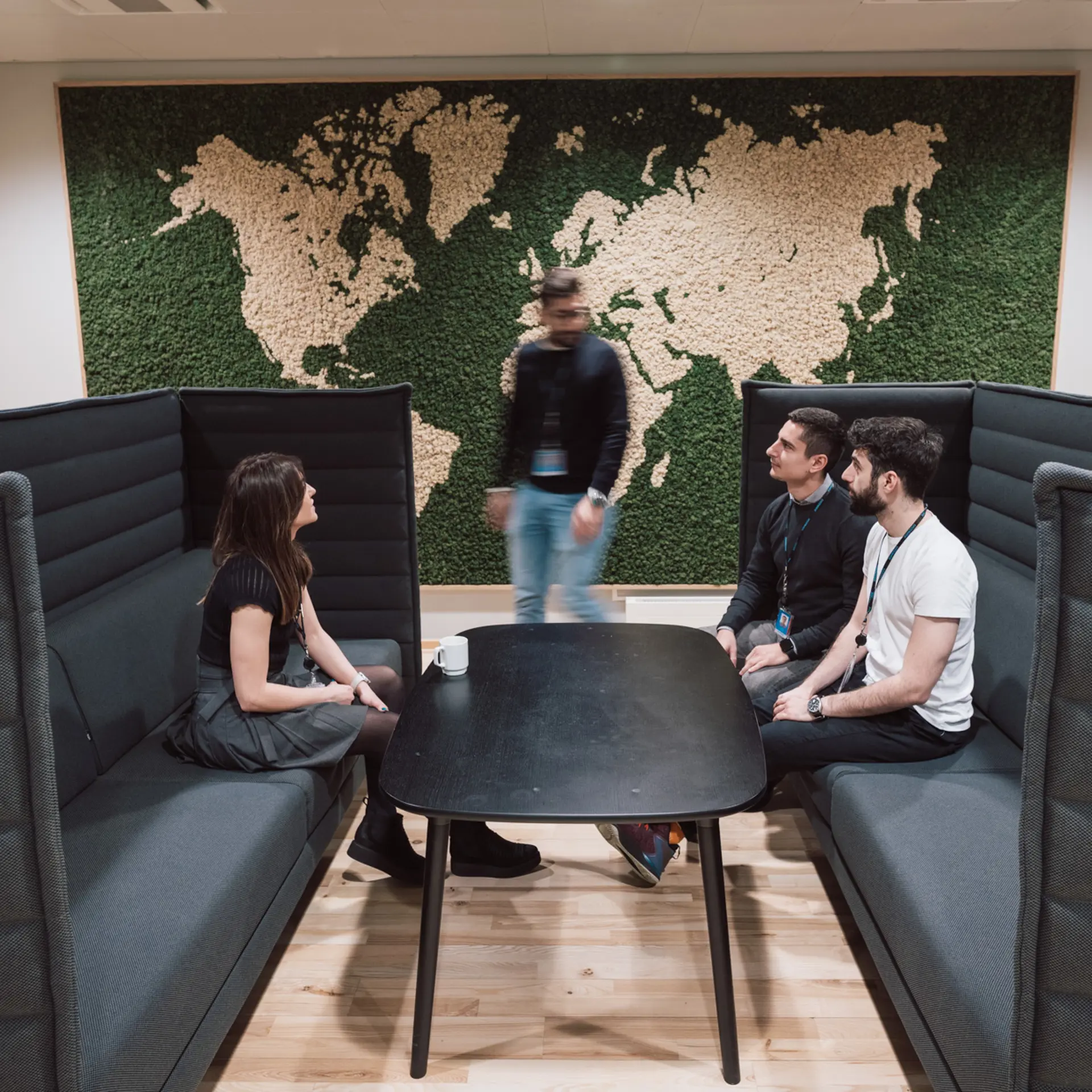 Systematic employees gathered for an informal meeting in a modern lounge area at Systematic HQ, with a world map wall feature in the background.