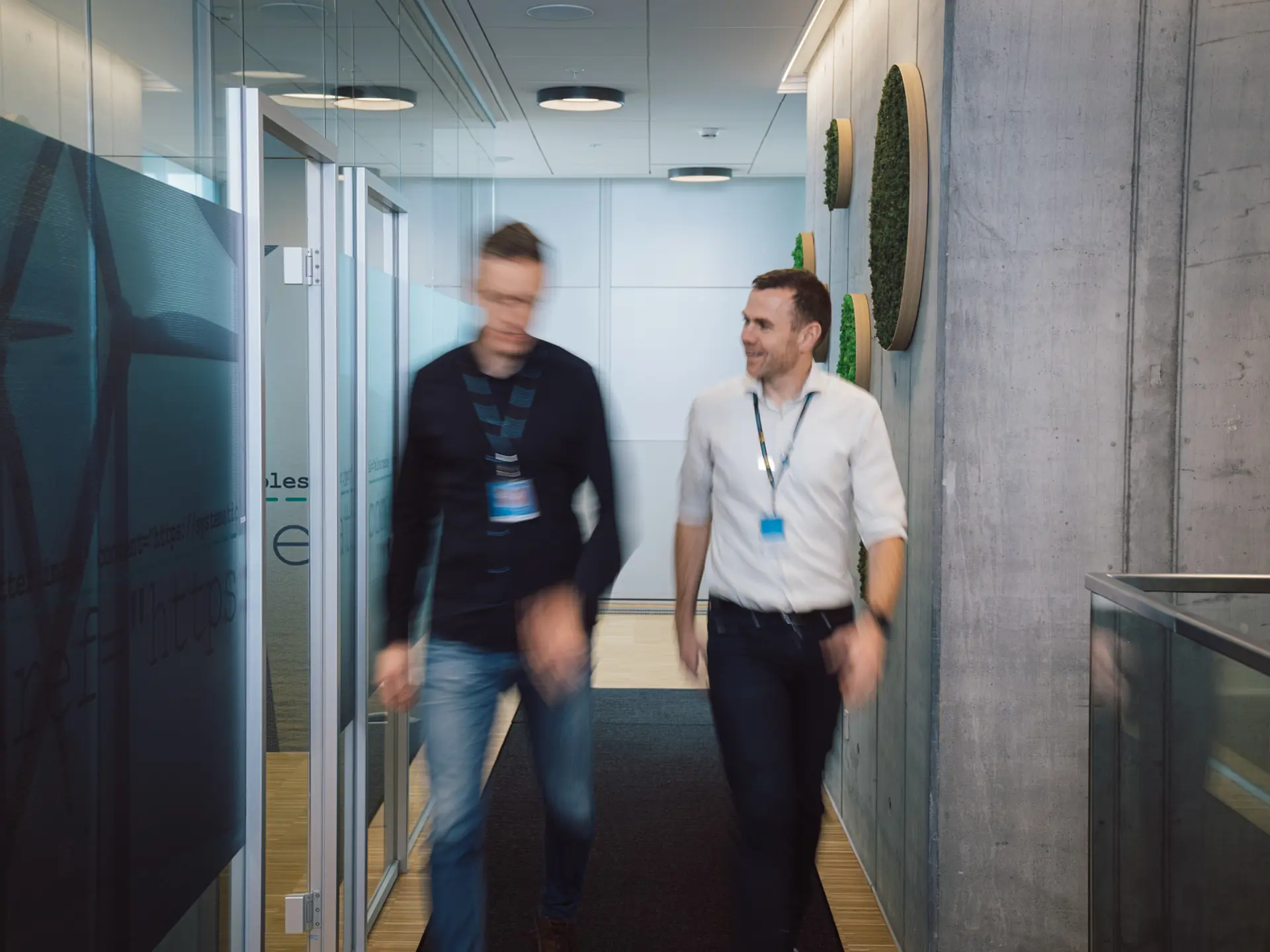 Systematic-anställda pratar medan de går genom en modern kontorskorridor med glasväggar och betongdetaljer.