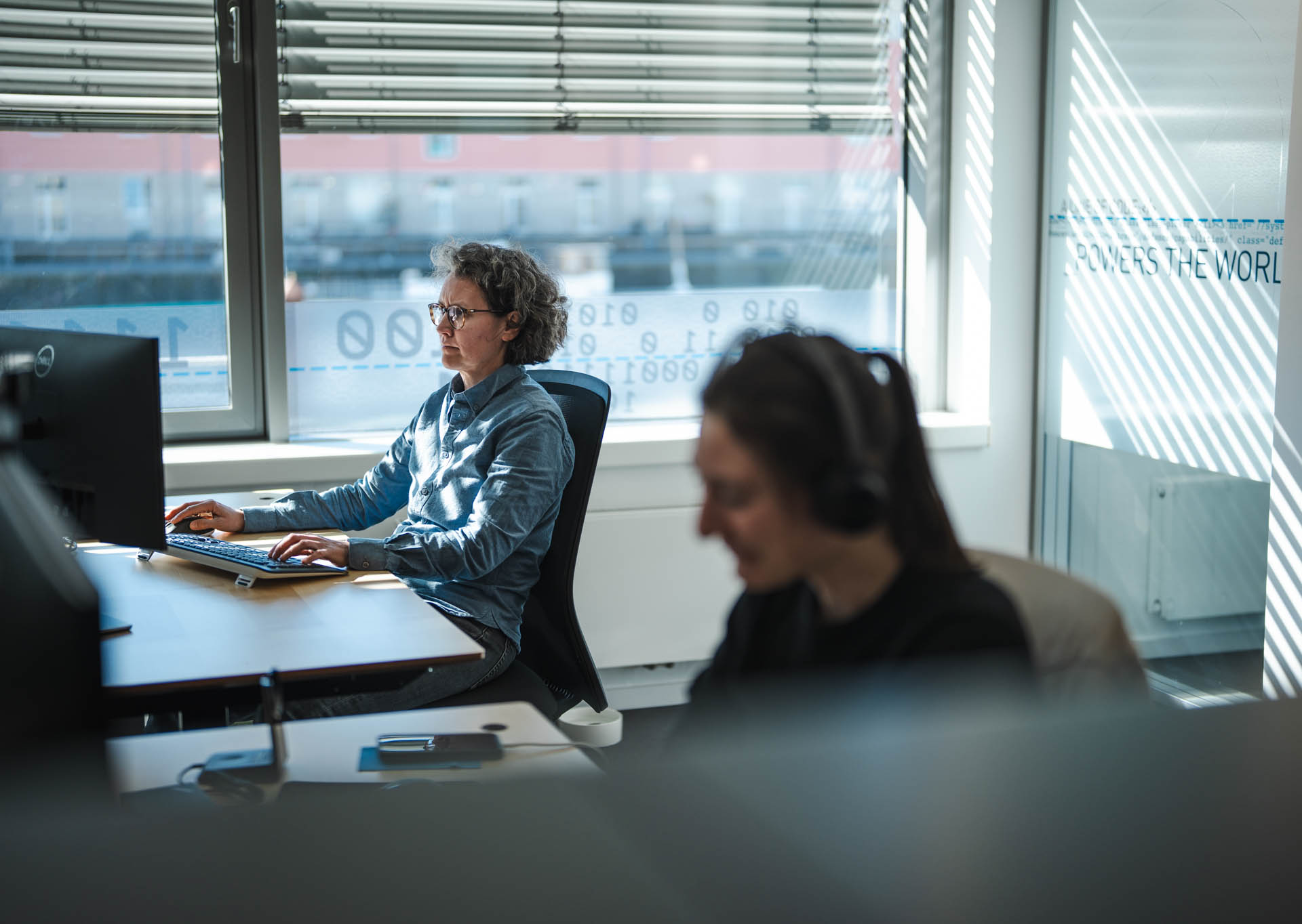 Two professionals working at computer workstations in a modern Systematic office with large windows and branded wall graphics.