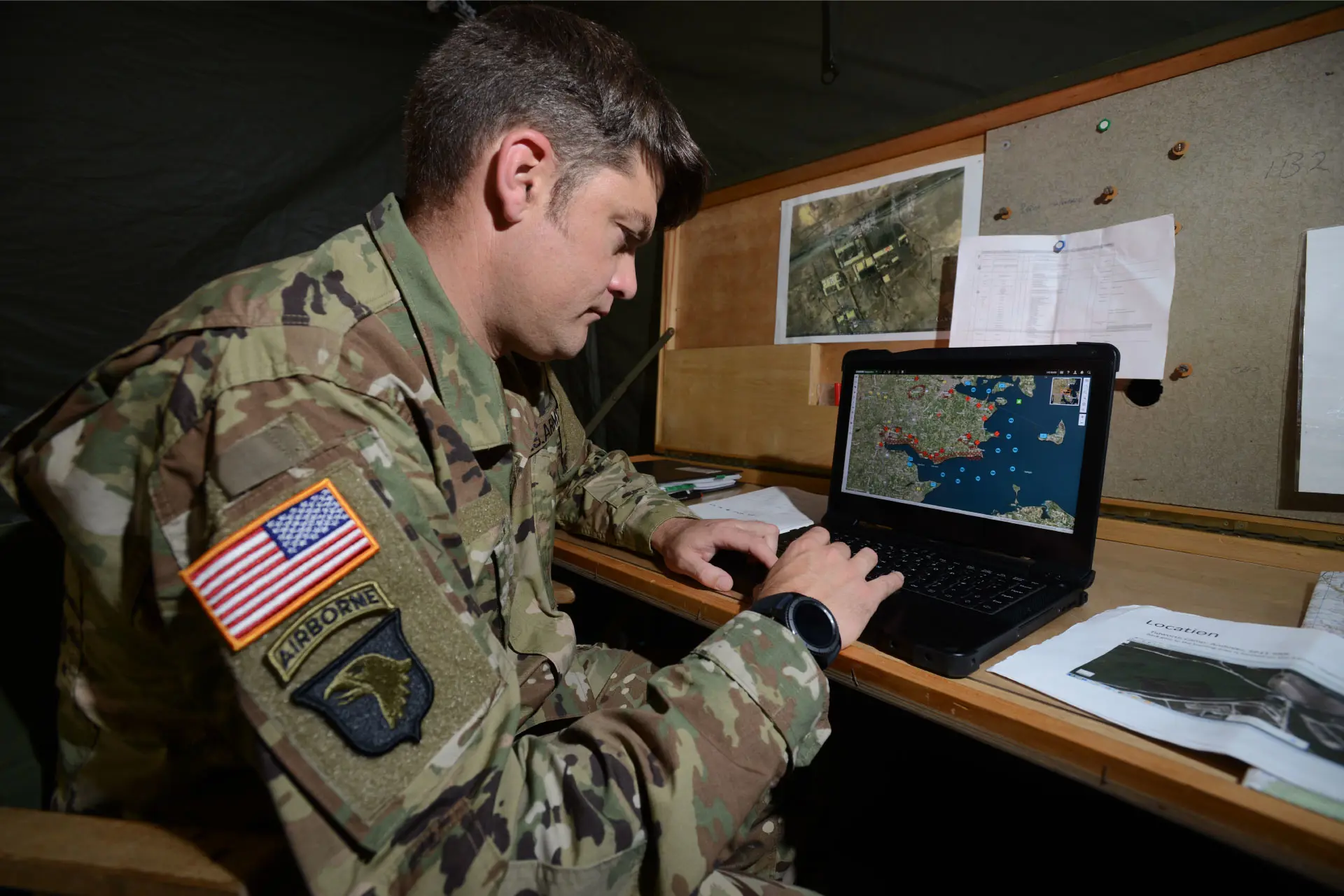 A U.S. Army airborne soldier in camouflage uniform analyzes a digital map on a laptop inside a military command tent.