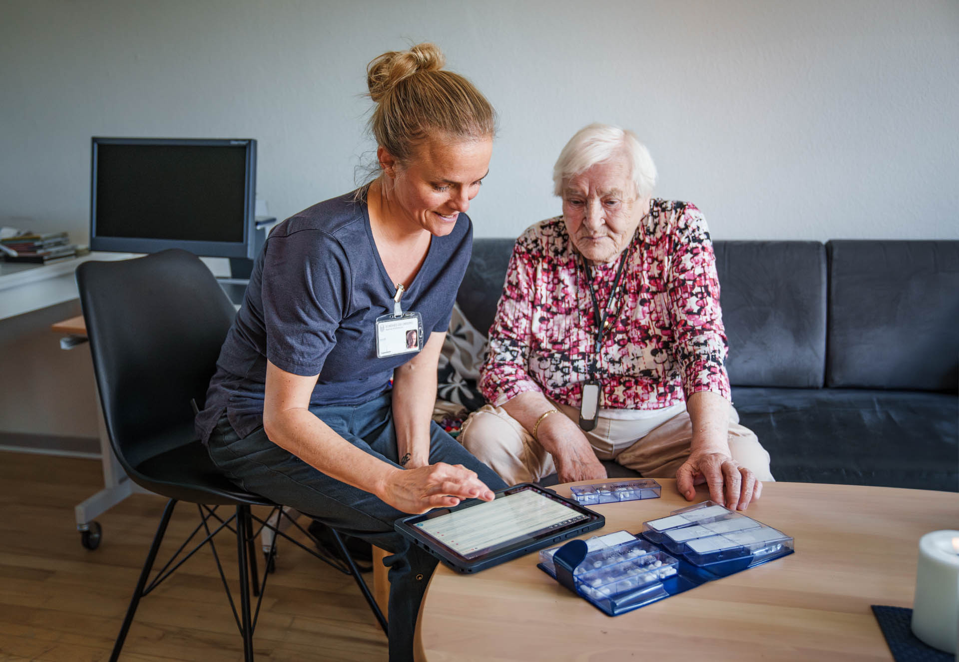 A caregiver helps an elderly woman manage her medication using a tablet, with pill organizers on the table. The woman wears a medical alert pendant.