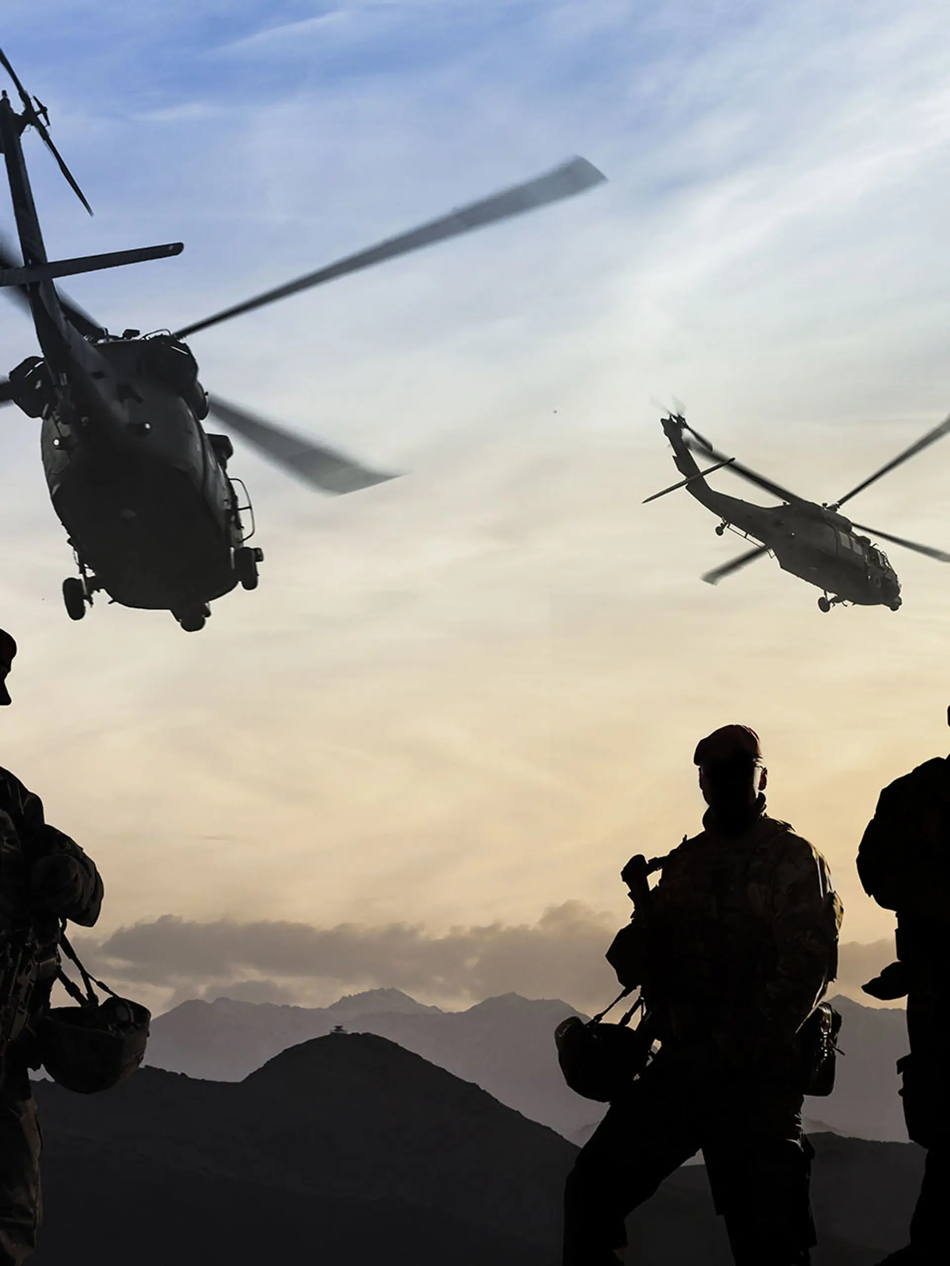 Soldiers silhouetted against the sunset with helicopters flying overhead during a military operation in the mountains.