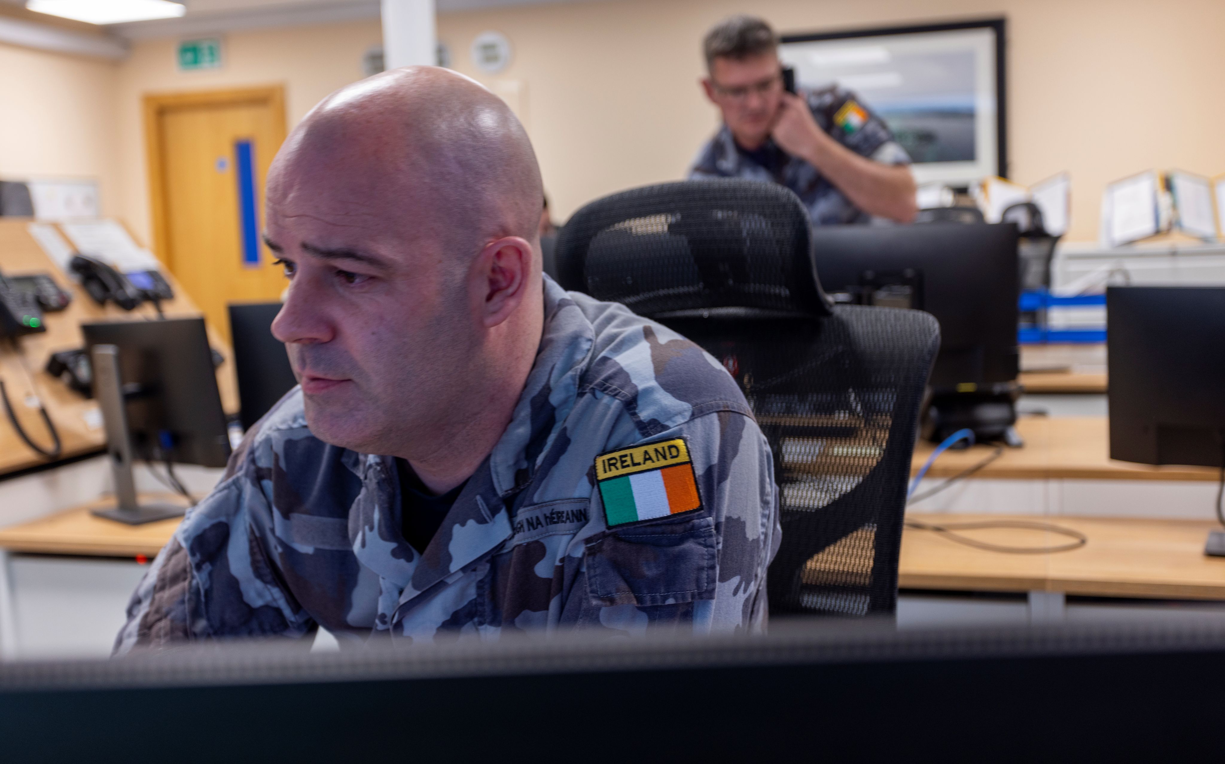 Irish military personnel working at a computer in an operations room, with a colleague on the phone in the background.