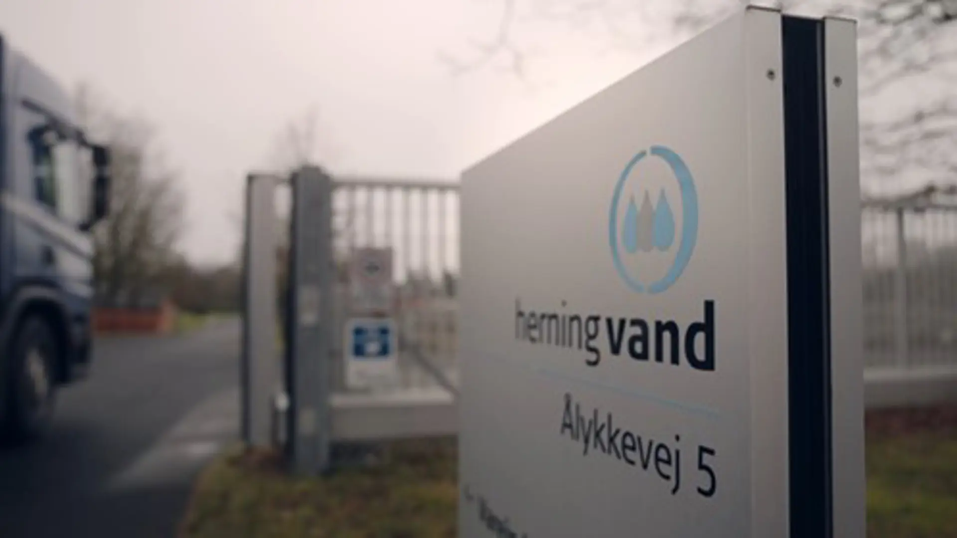 Entrance sign for Herning Vand at Ålykkevej 5, next to a security gate and driveway with a truck entering the facility on a cloudy day.