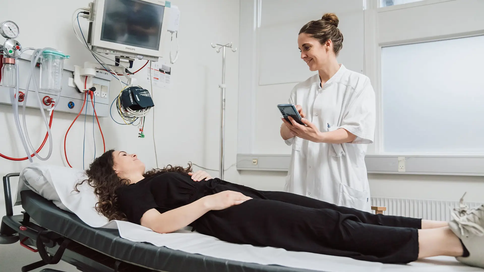 Healthcare professional using mobile device while monitoring patient on medical examination table in modern clinical room with diagnostic equipment