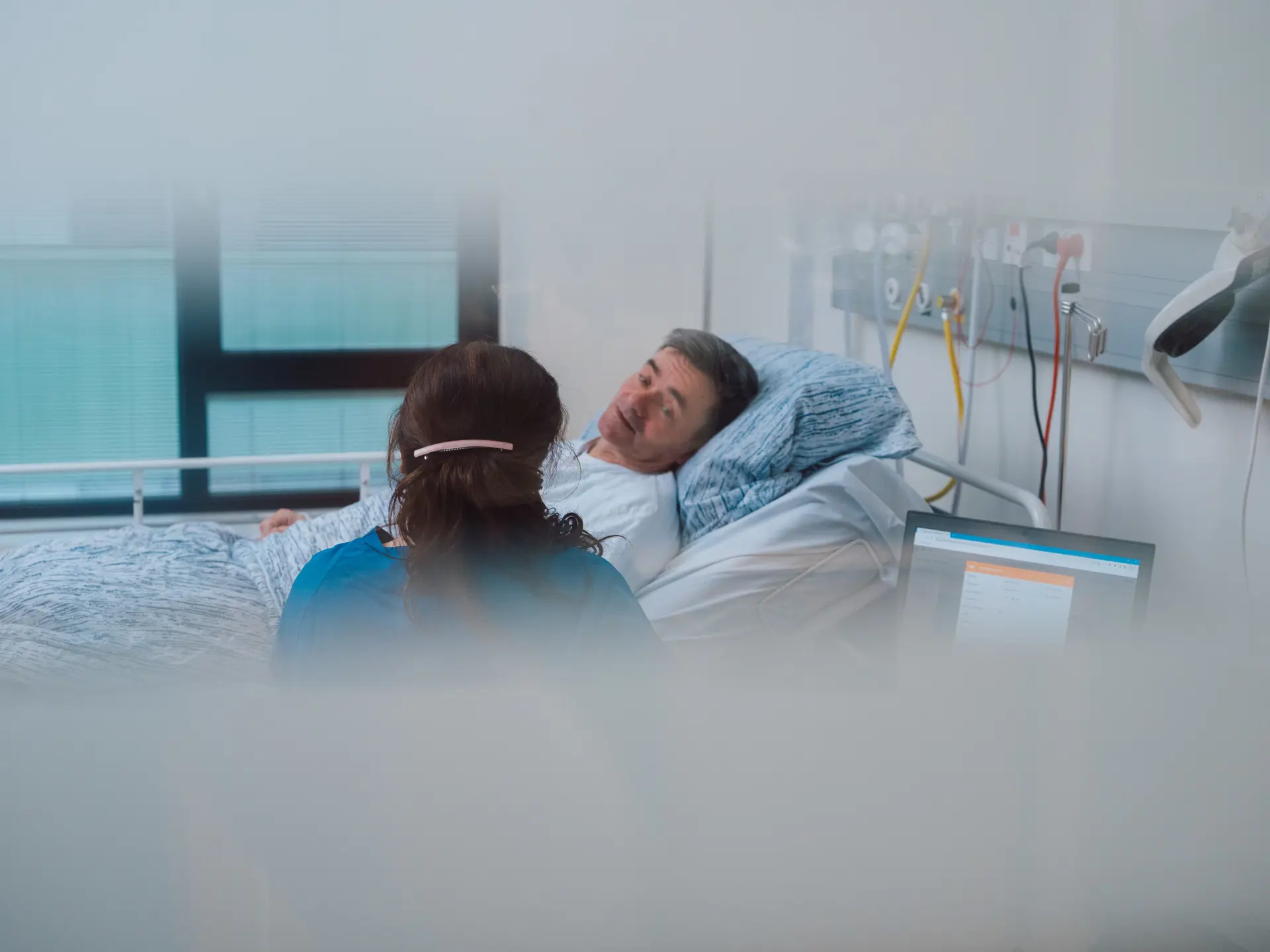 A nurse in blue scrubs sits beside a male patient in a hospital bed, engaging in conversation in a modern medical facility.