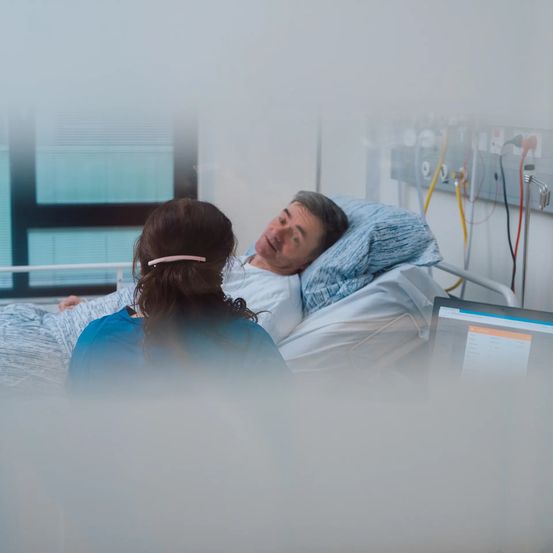 A nurse in blue scrubs sits beside a male patient in a hospital bed, engaging in conversation in a modern medical facility.