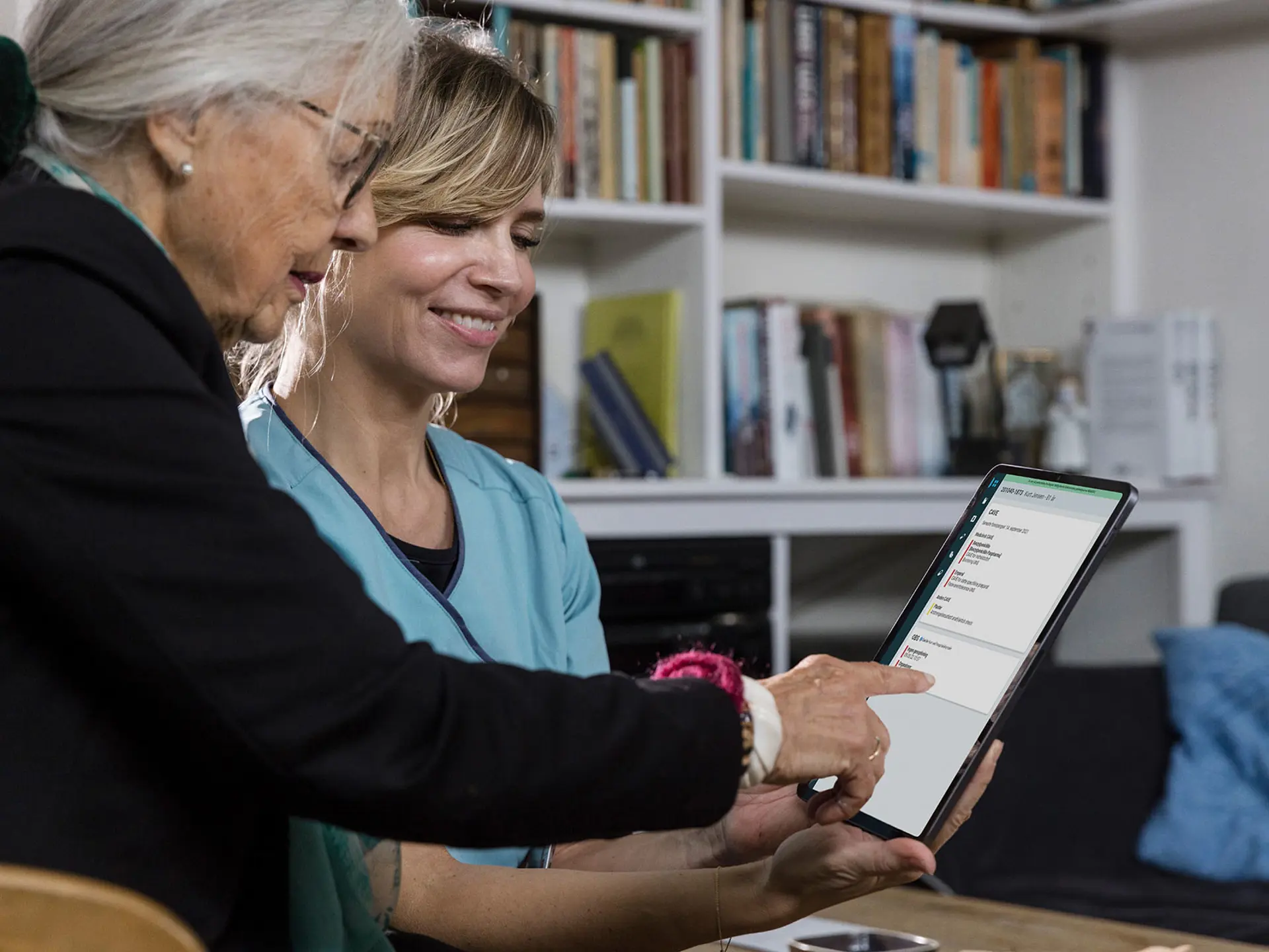 Smilende sundhedsprofessionel, der hjælper en ældre kvinde, mens de sammen bruger en tablet i en hyggelig dagligstue med bogreoler i baggrunden.