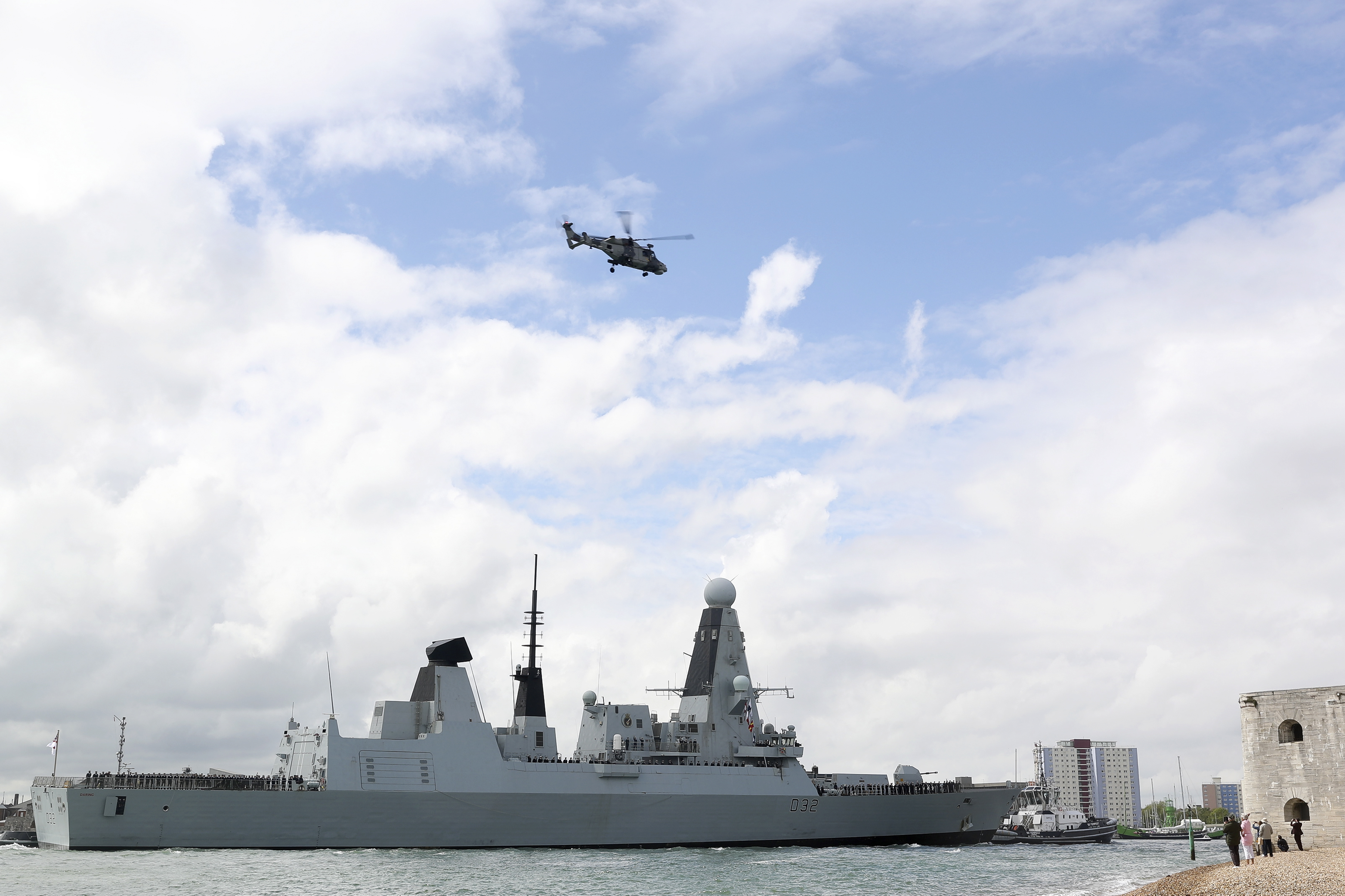 A naval battleship sails near the coast as a military helicopter flies overhead under a partly cloudy sky