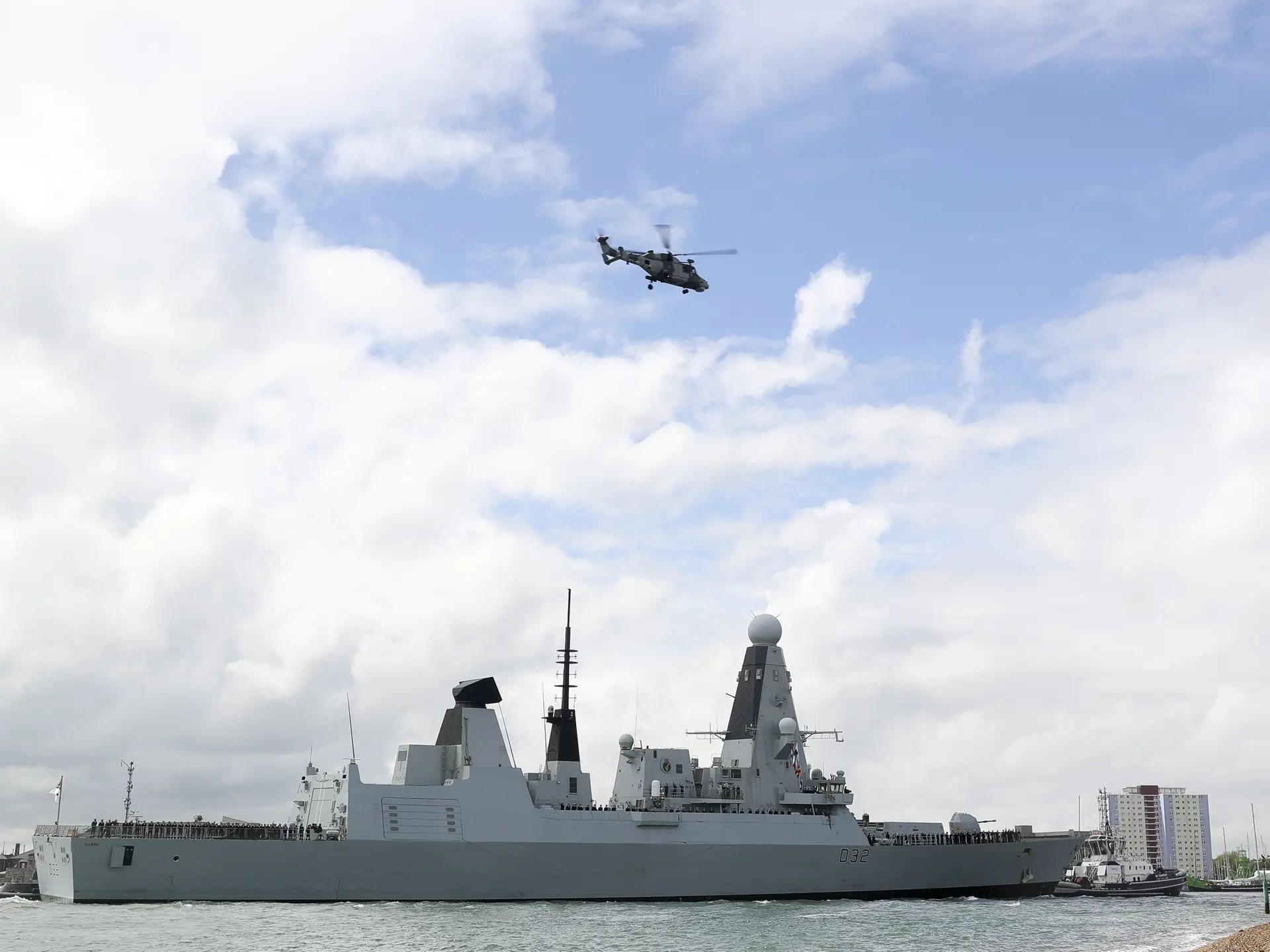 A naval battleship sails near the coast as a military helicopter flies overhead under a partly cloudy sky