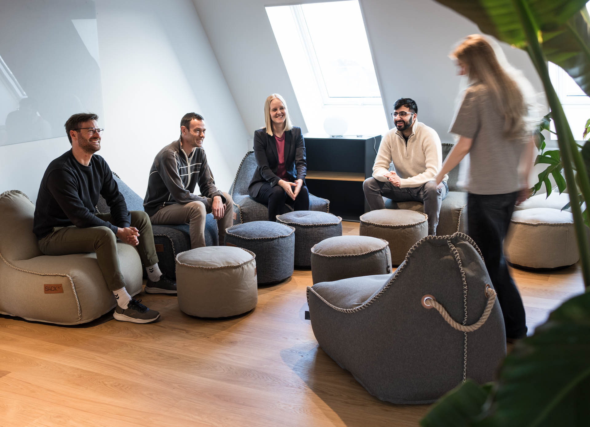Four smiling colleagues sit on bean bag chairs in a bright attic lounge at Systematic’s Copenhagen office as a fifth person walks past.
