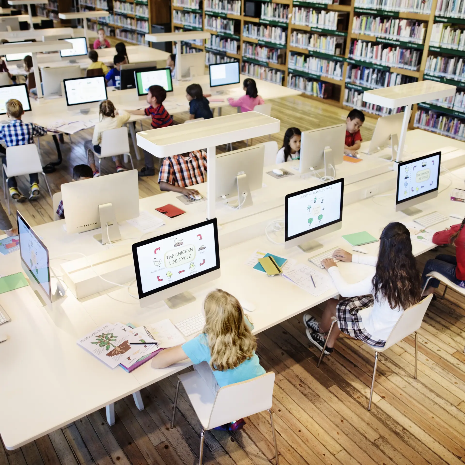 Børn arbejder ved stationære computere i et moderne bibliotekslokale, mens en lærer hjælper dem, omgivet af bogreoler i baggrunden.