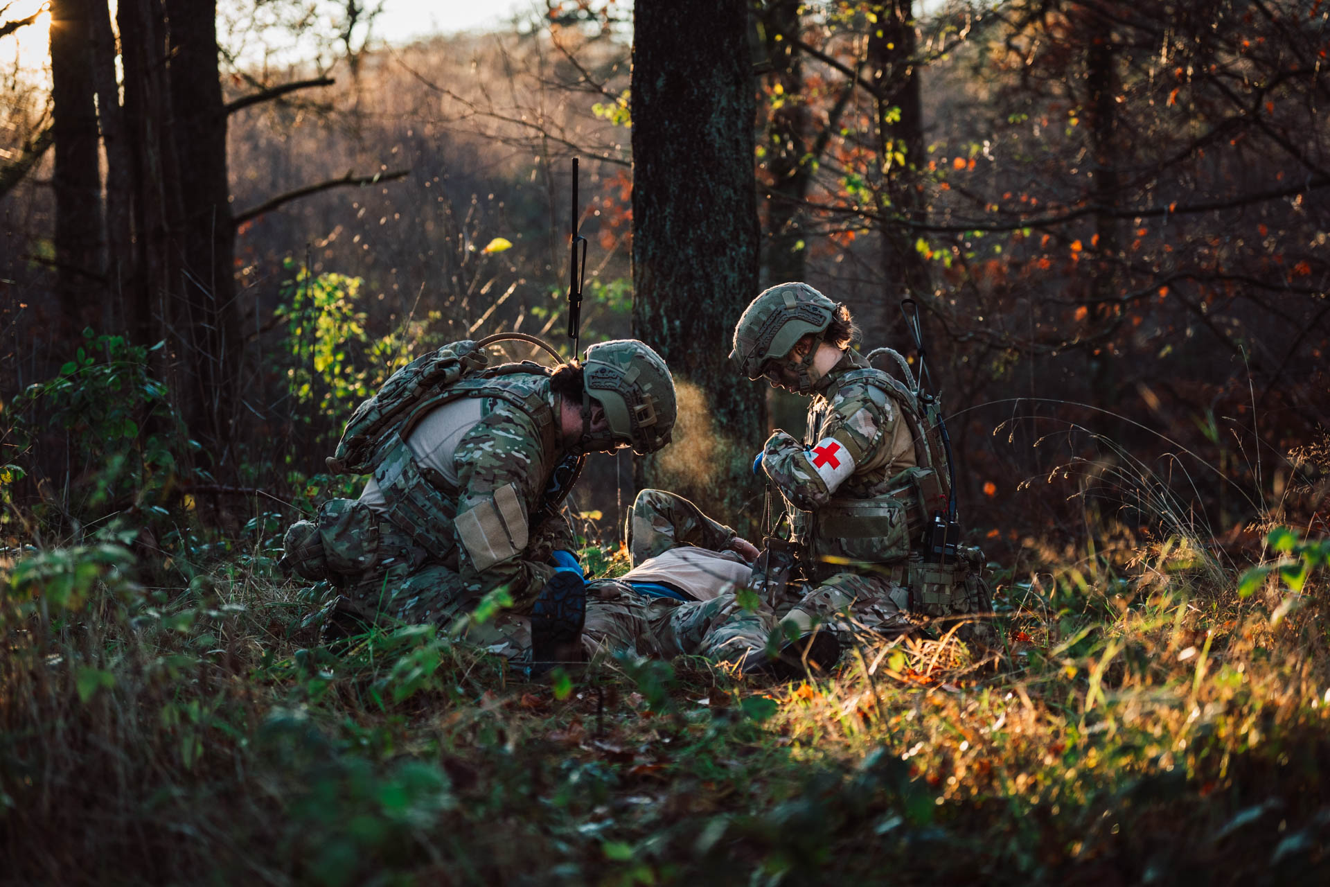 Two military medics in camouflage uniforms provide tactical first aid to an injured soldier lying on the ground during a woodland training exercise.