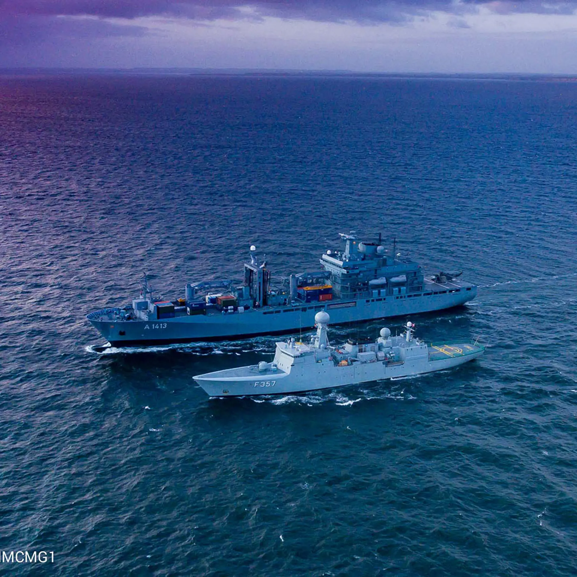 NATO warships A1413 and F357 sail side by side on open water at sunset, with dramatic purple and blue tones in the sky and sea.