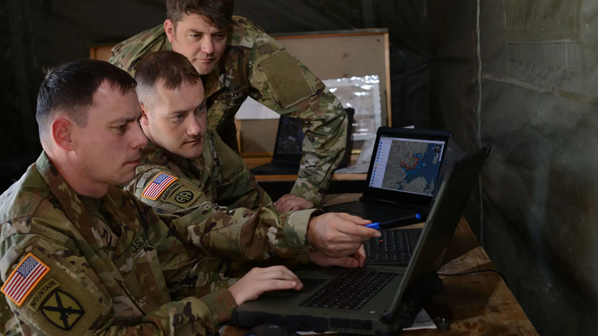 Soldiers inside a field tent reviewing a digital battlefield map on a rugged laptop, with a commander pointing at the screen during operational planning.