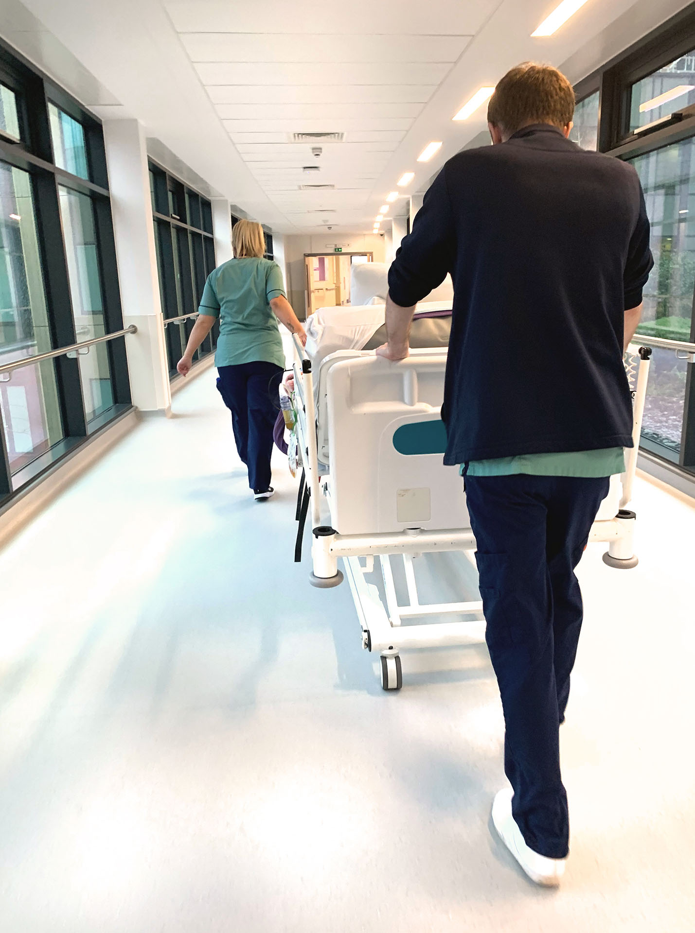 Two hospital staff members transporting empty beds along a bright hospital corridor.