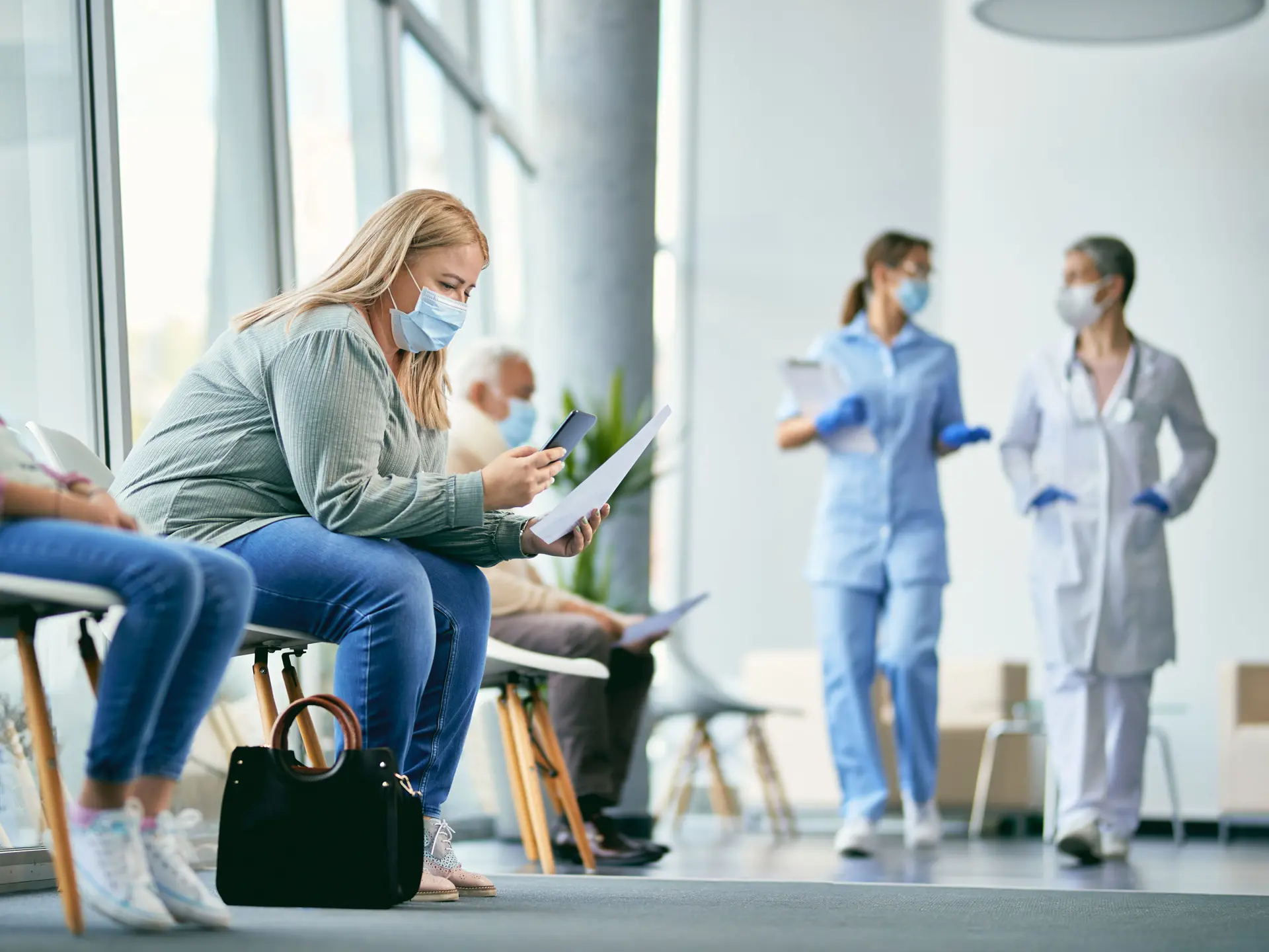 Female patient examining medical paperwork and using mobile phone while sitting in hallway at the hospital