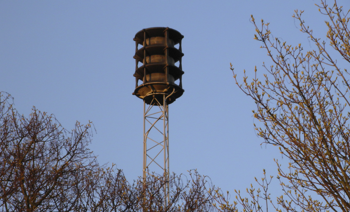 Outdoor warning siren mounted on a steel tower against a clear sky, part of Denmark's national siren warning system.