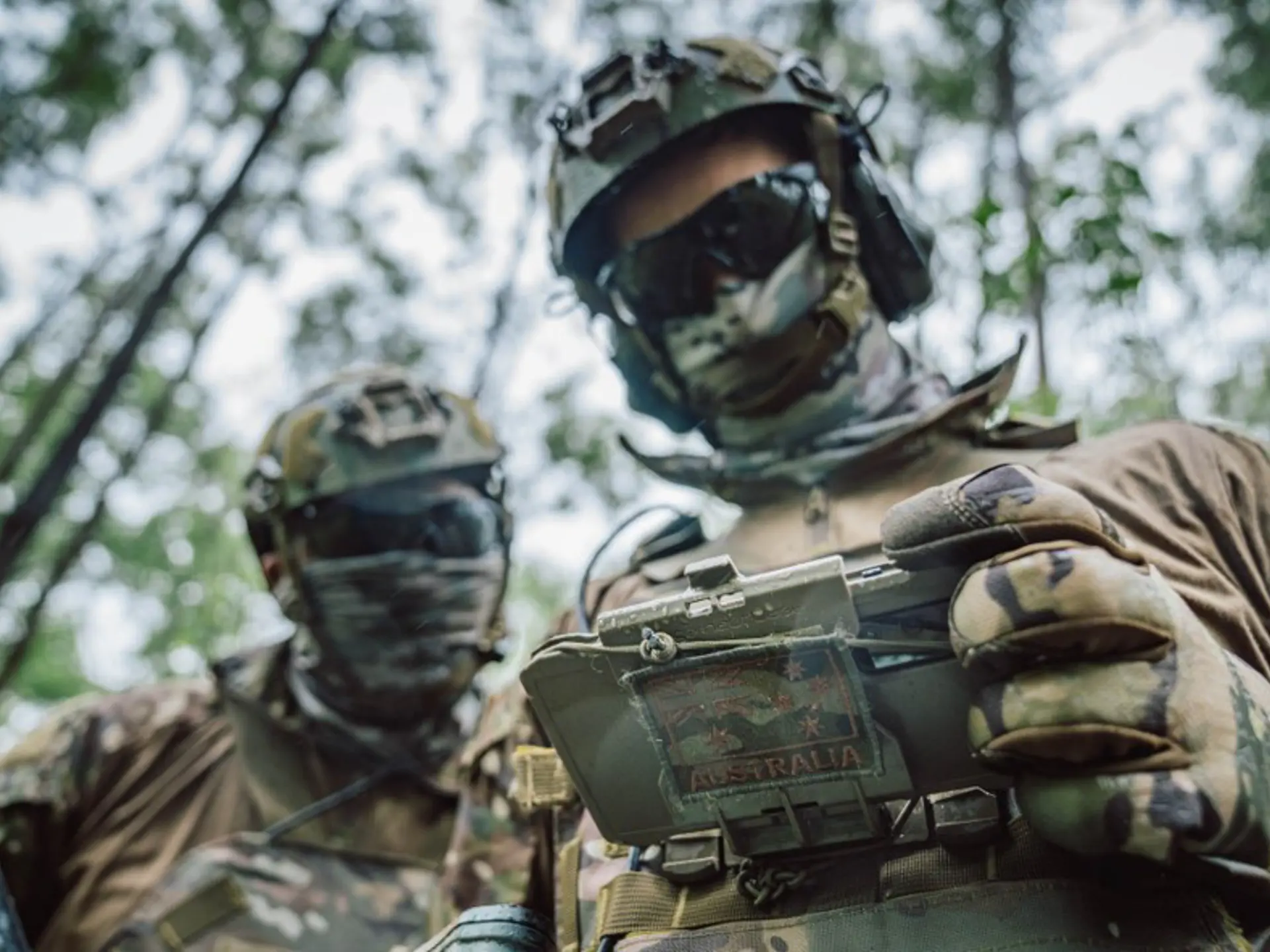 Two military personnel in camouflage and helmets in a forest, one holding a handheld device with an Australian flag patch.