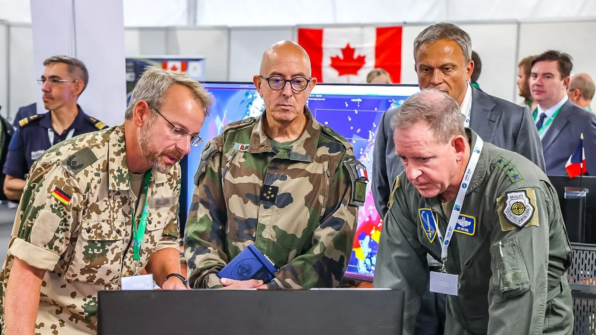 Military officers from different nations examine a computer screen at a defence exhibition, with a Canadian flag in the background.