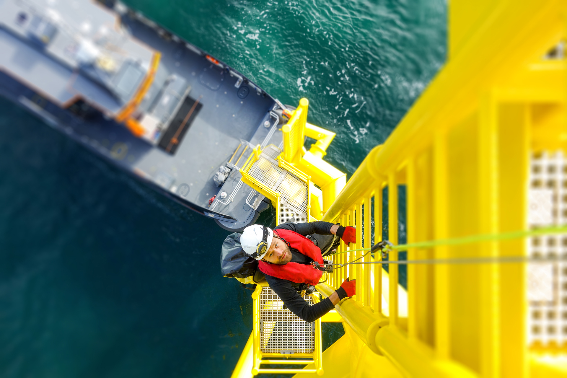 Offshore technician wearing safety gear climbing a yellow ladder structure above the water with a service vessel below.