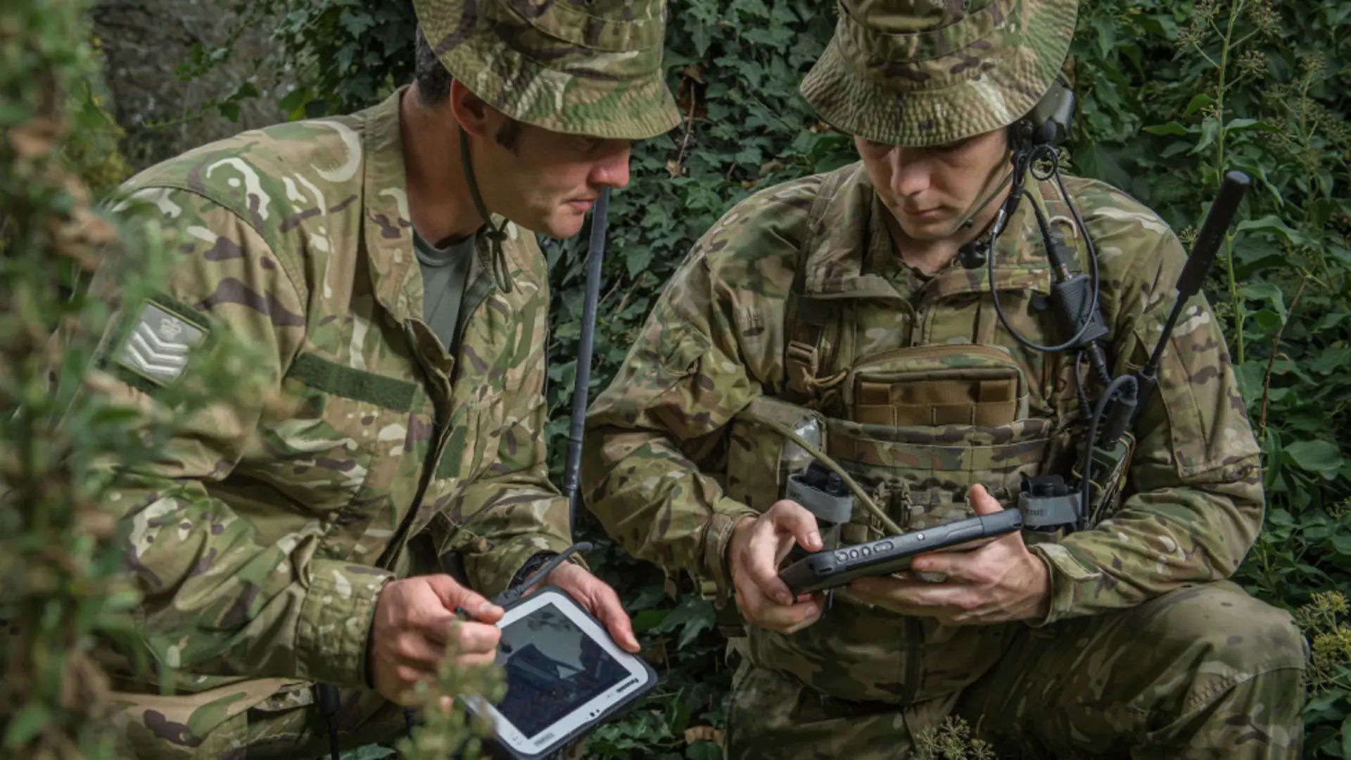 Soldiers in camouflage use military tablets for communication in a dense, green environment.