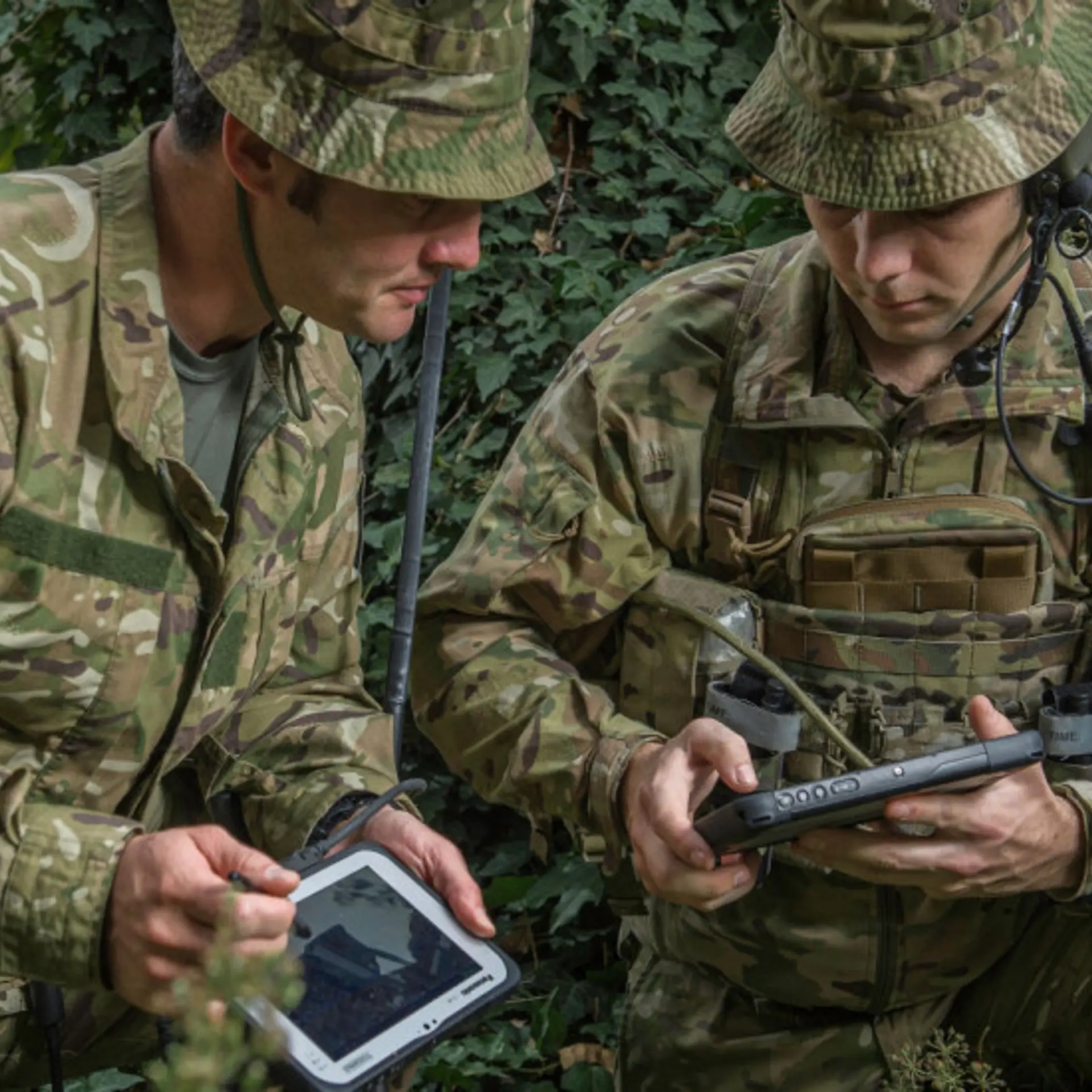 Soldiers in camouflage use military tablets for communication in a dense, green environment.