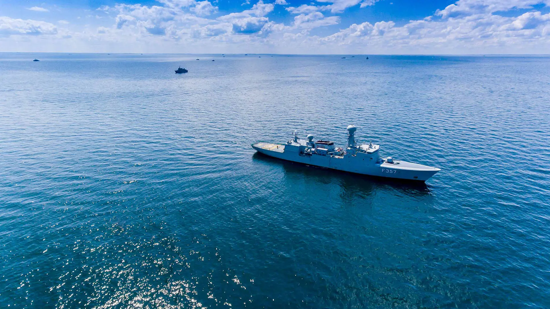 Ein Marineschiff fährt durch ruhige, offene Gewässer unter einem strahlend blauen Himmel, mit weiteren Schiffen in der Ferne.
