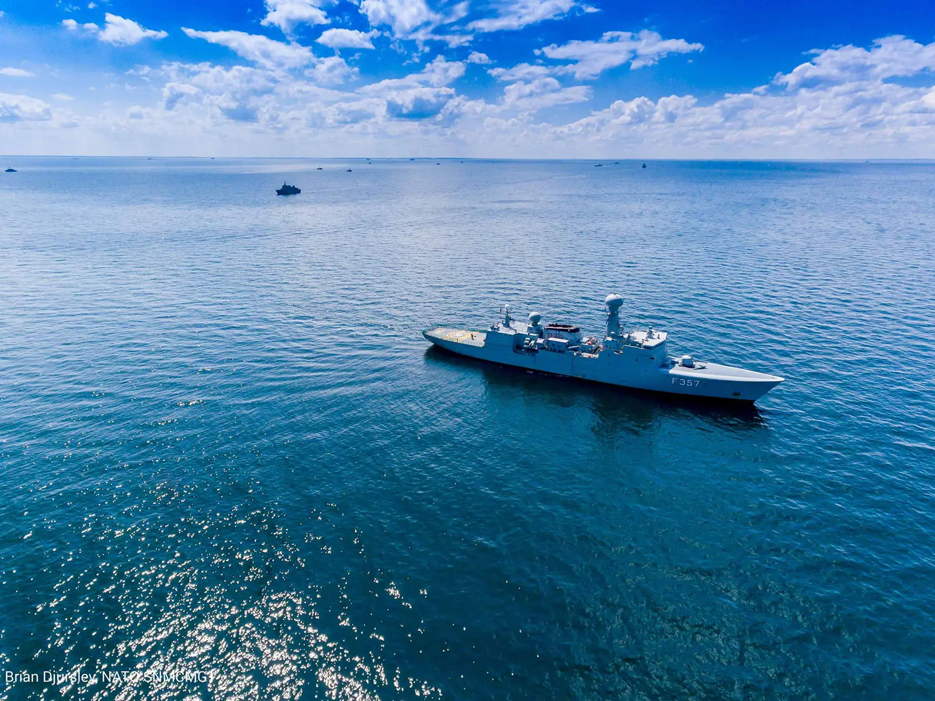A naval vessel sails through calm open waters under a bright blue sky, with other ships visible in the distance.