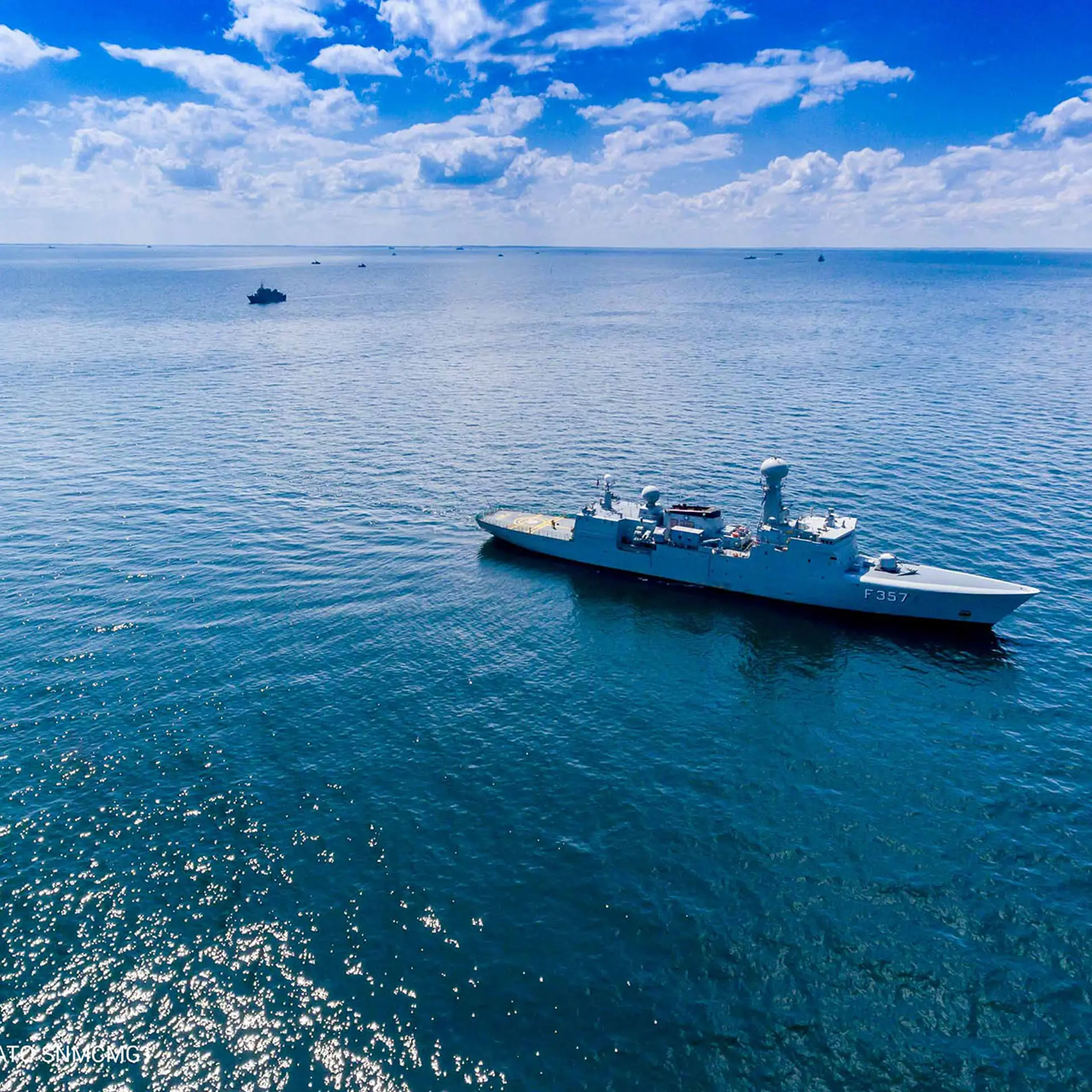 A naval vessel sails through calm open waters under a bright blue sky, with other ships visible in the distance.