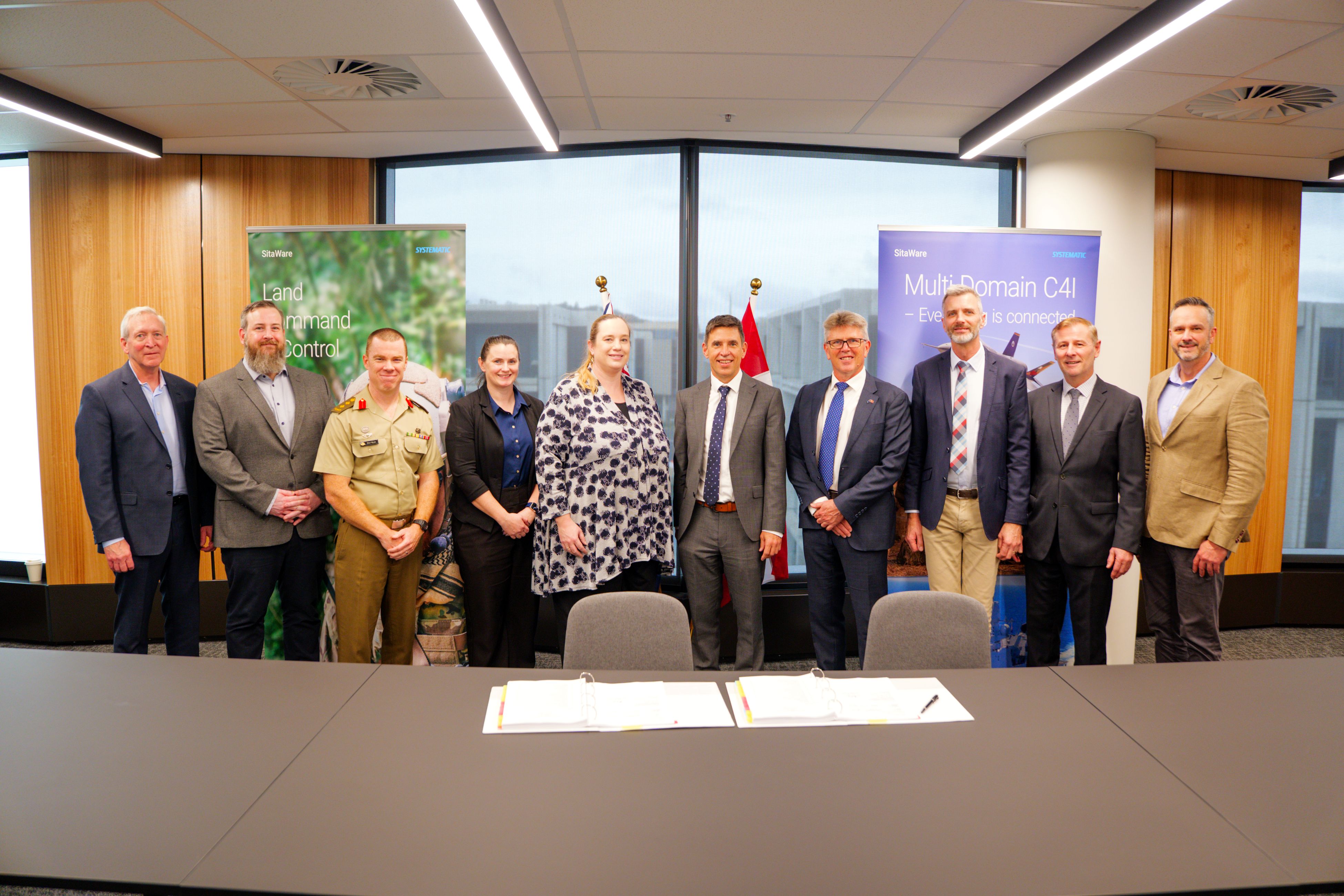 Group of Systematic and defence representatives, including military personnel, posing after signing the Land 4140 contract, with product banners in the background.