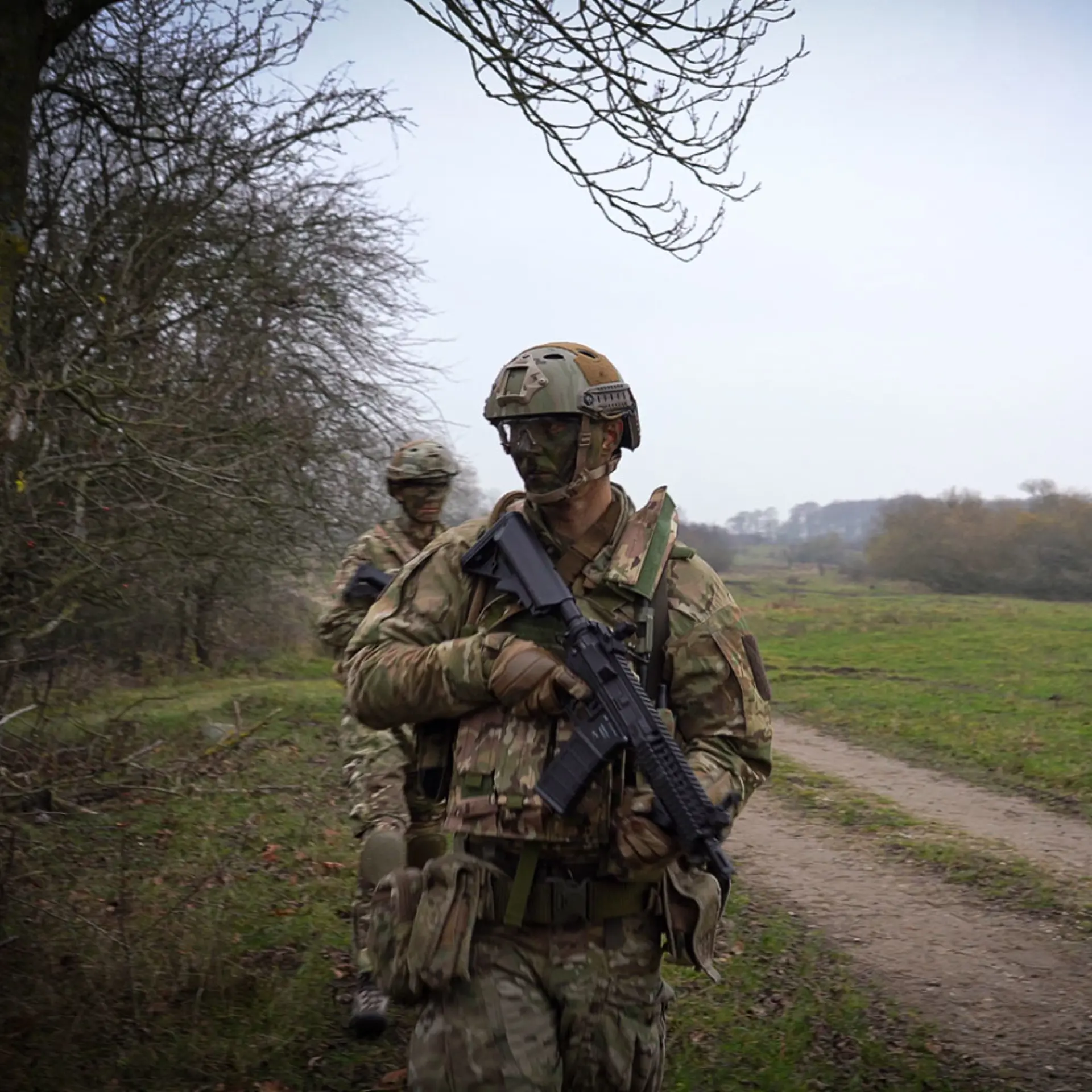 Two soldiers in camouflage gear with rifles patrol a rural dirt path next to a forest during a military training exercise