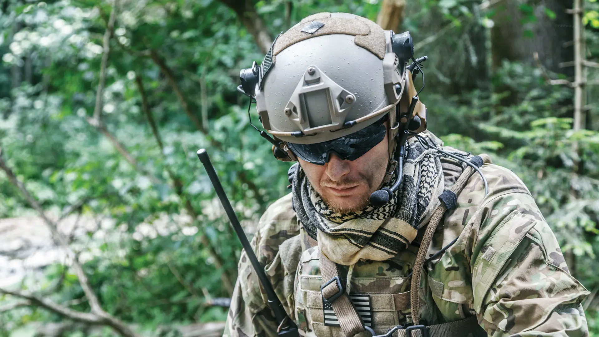 A soldier in camouflage tactical gear and a helmet with a mounted camera, communicating via a radio and checking a device in a dense forest