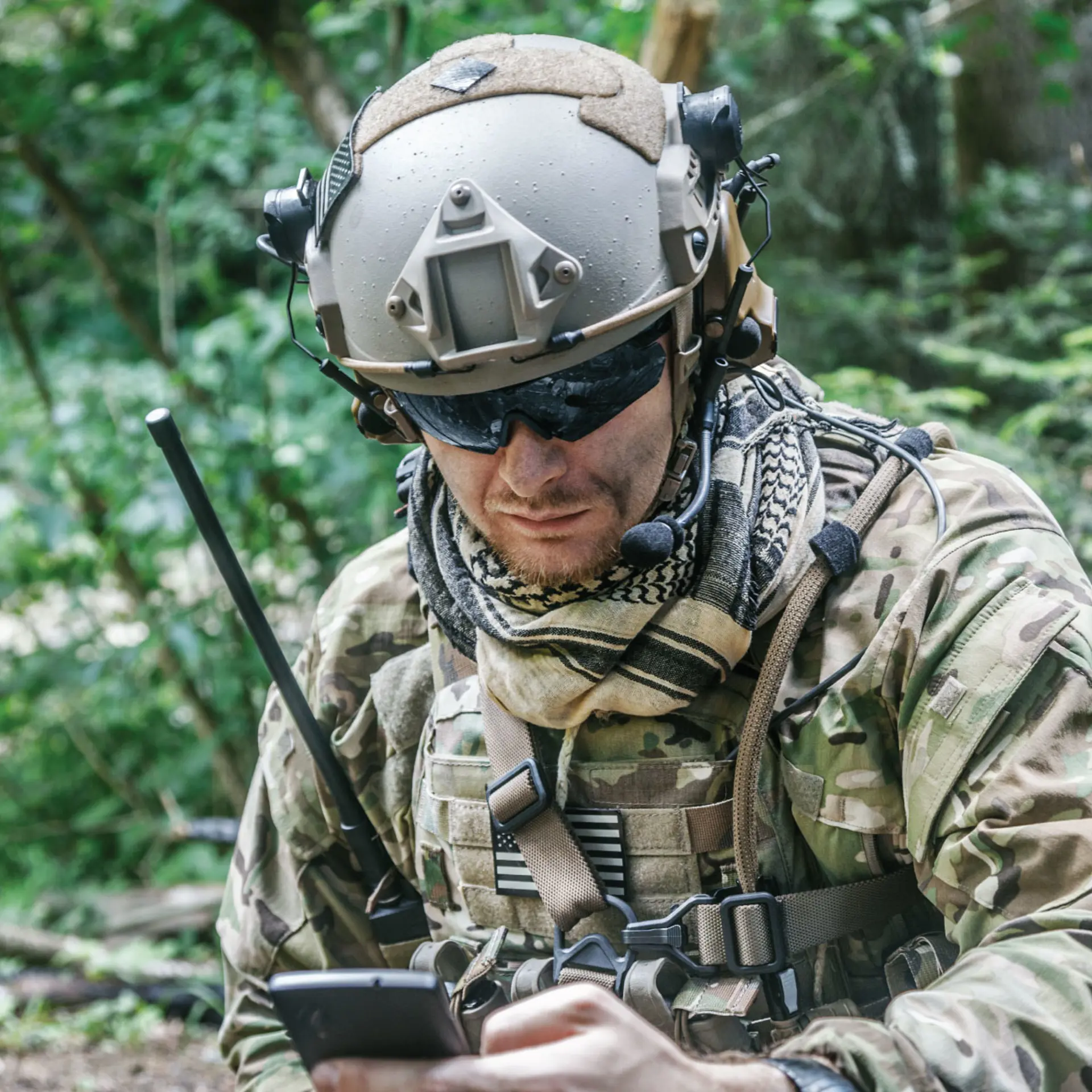 A soldier in camouflage tactical gear and a helmet with a mounted camera, communicating via a radio and checking a device in a dense forest