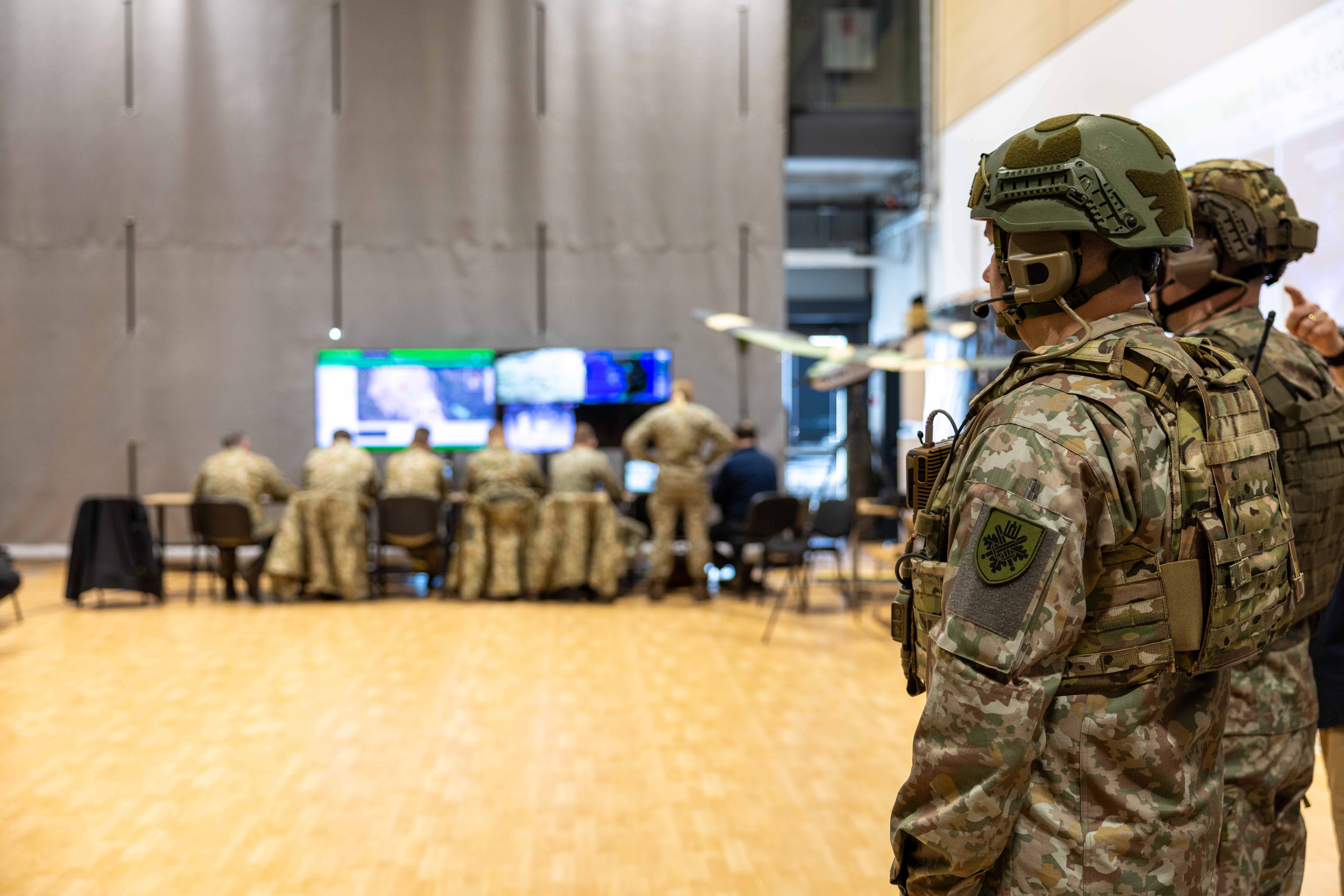 Armed military personnel in combat gear and helmets observe a tactical briefing with digital displays during Lithuania's War Herald 2026 exercise in an operations center.