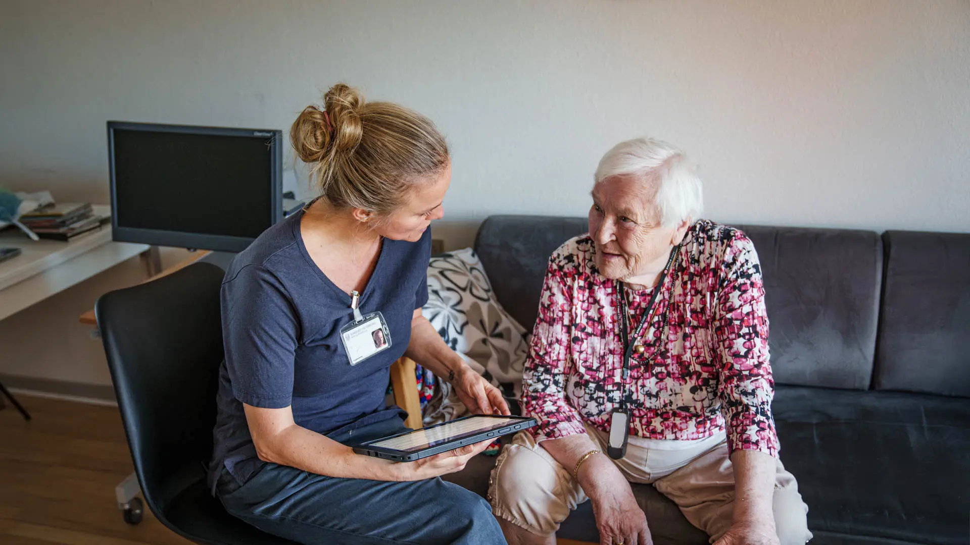 A caregiver in a navy-blue uniform shows a tablet to an elderly woman in a floral blouse, seated on a black sofa in a well-lit living room.